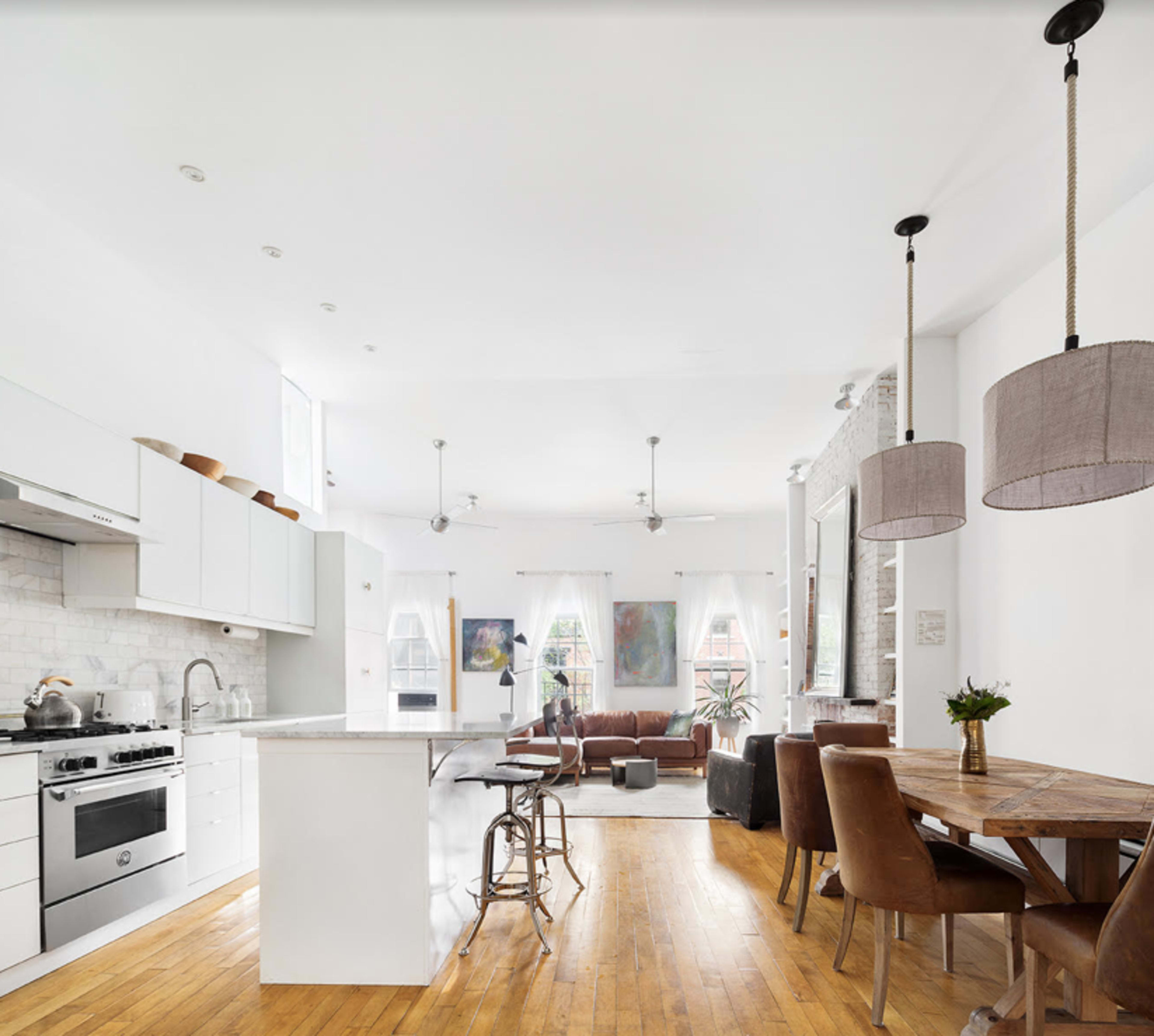 A modern kitchen and dining area with white cabinetry, a central island, a wooden dining table, and a brown leather sofa.