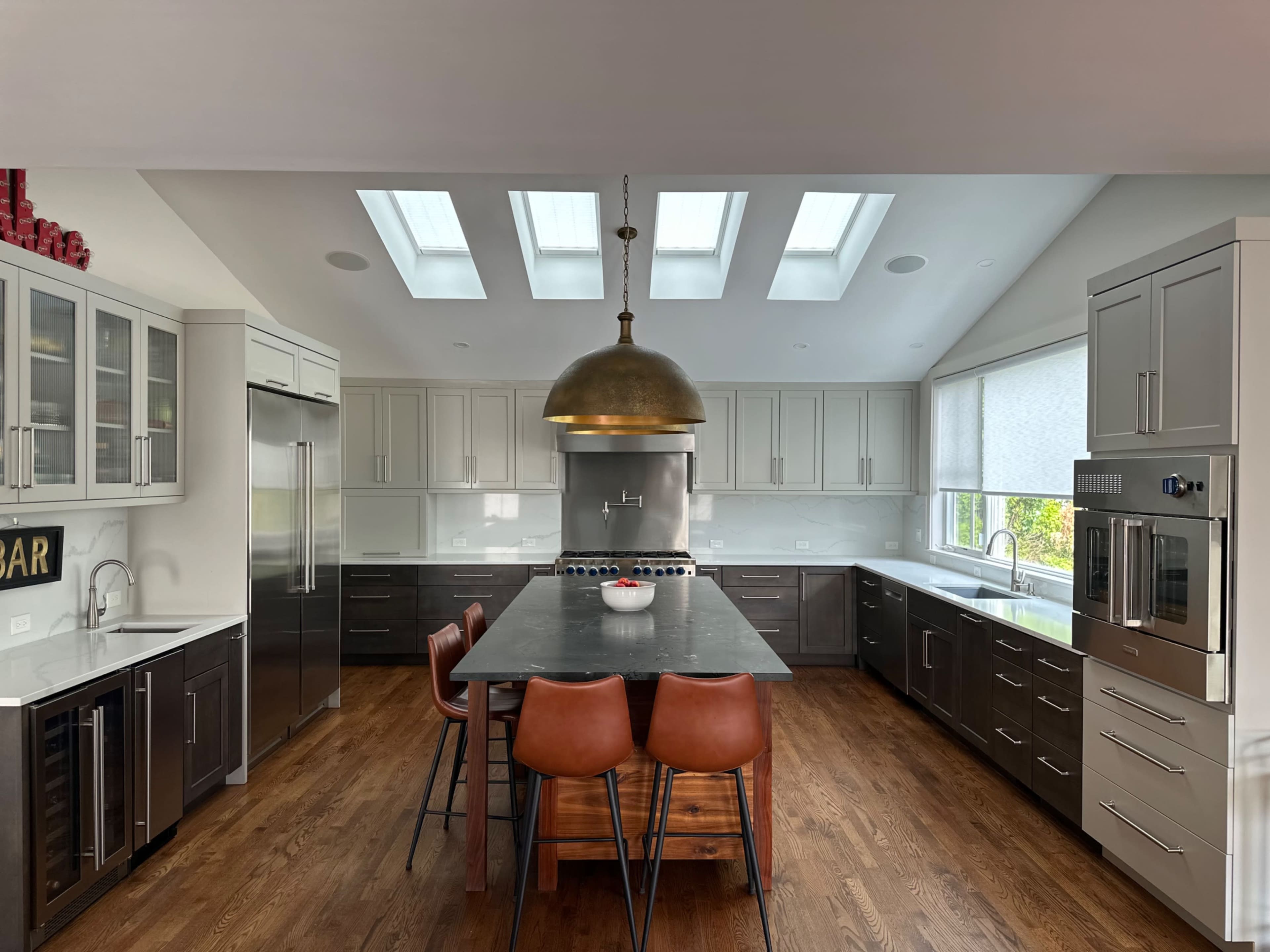 A modern kitchen features a large central island with brown bar stools, stainless steel appliances, and multiple skylights providing natural light.