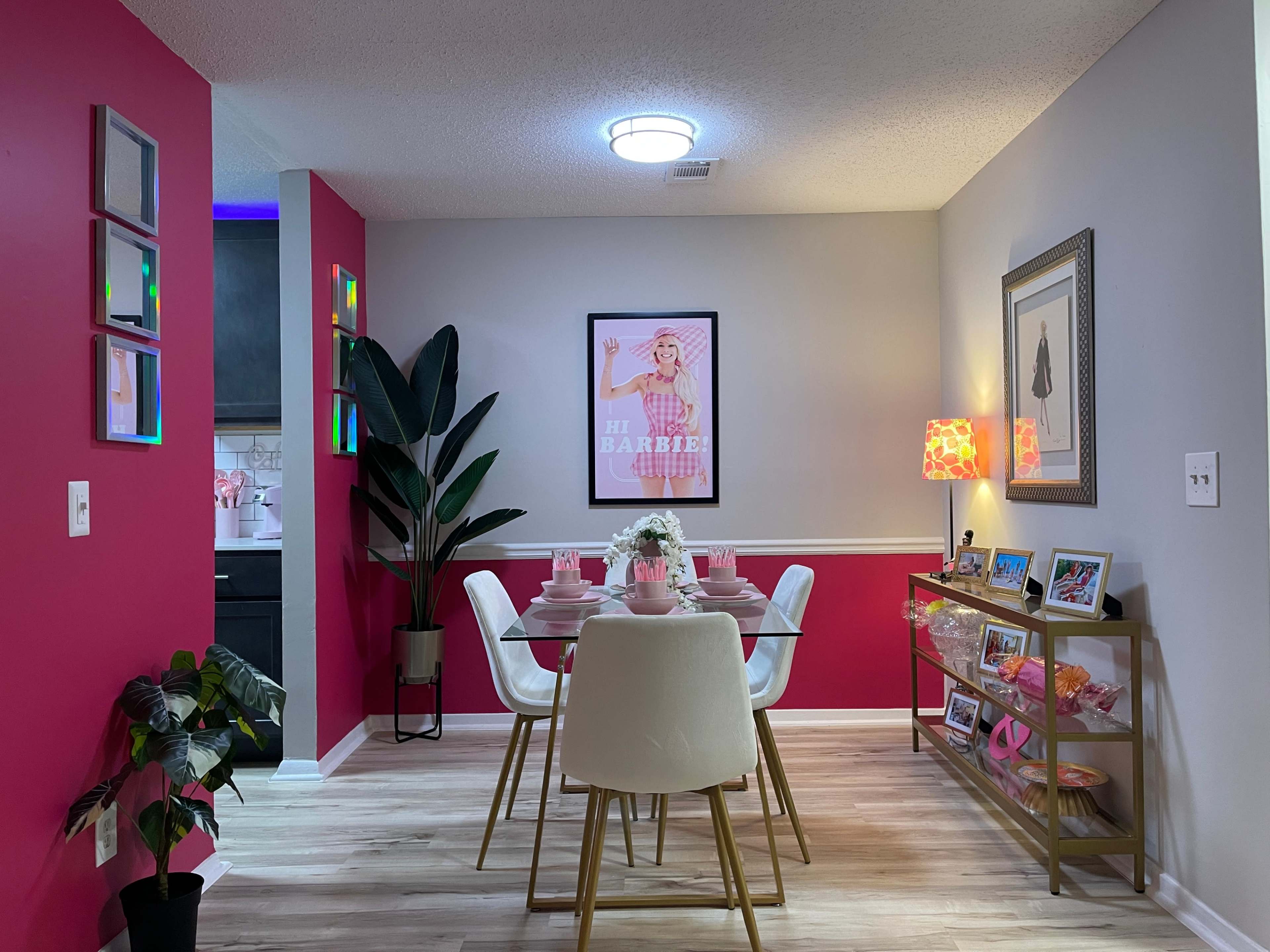 A dining area features a pink accent wall, a round table set for four, and framed artwork, with a potted plant positioned nearby.