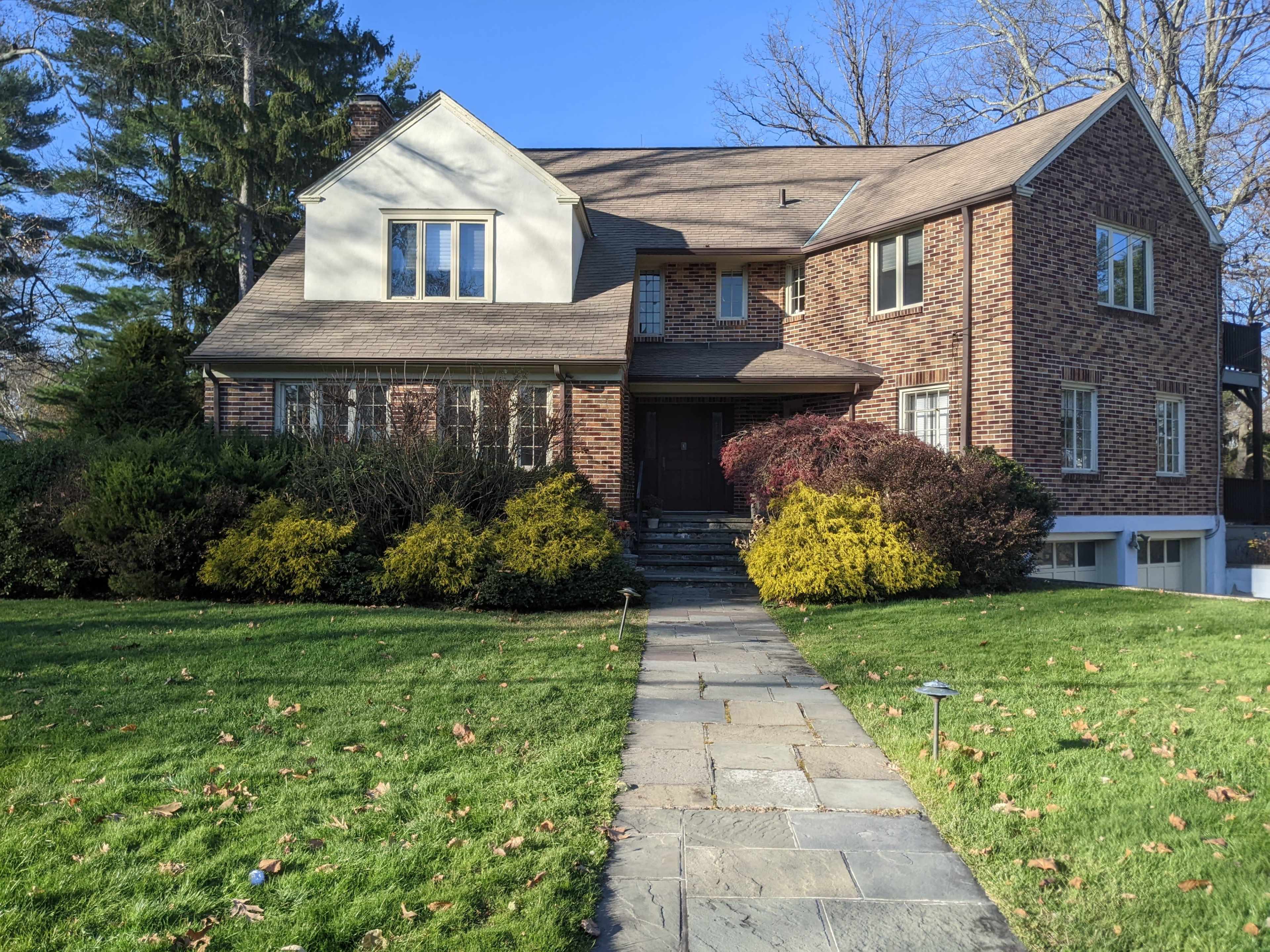 A large brick house with multiple windows and a front garden featuring shrubs and a stone walkway leads to the entrance.