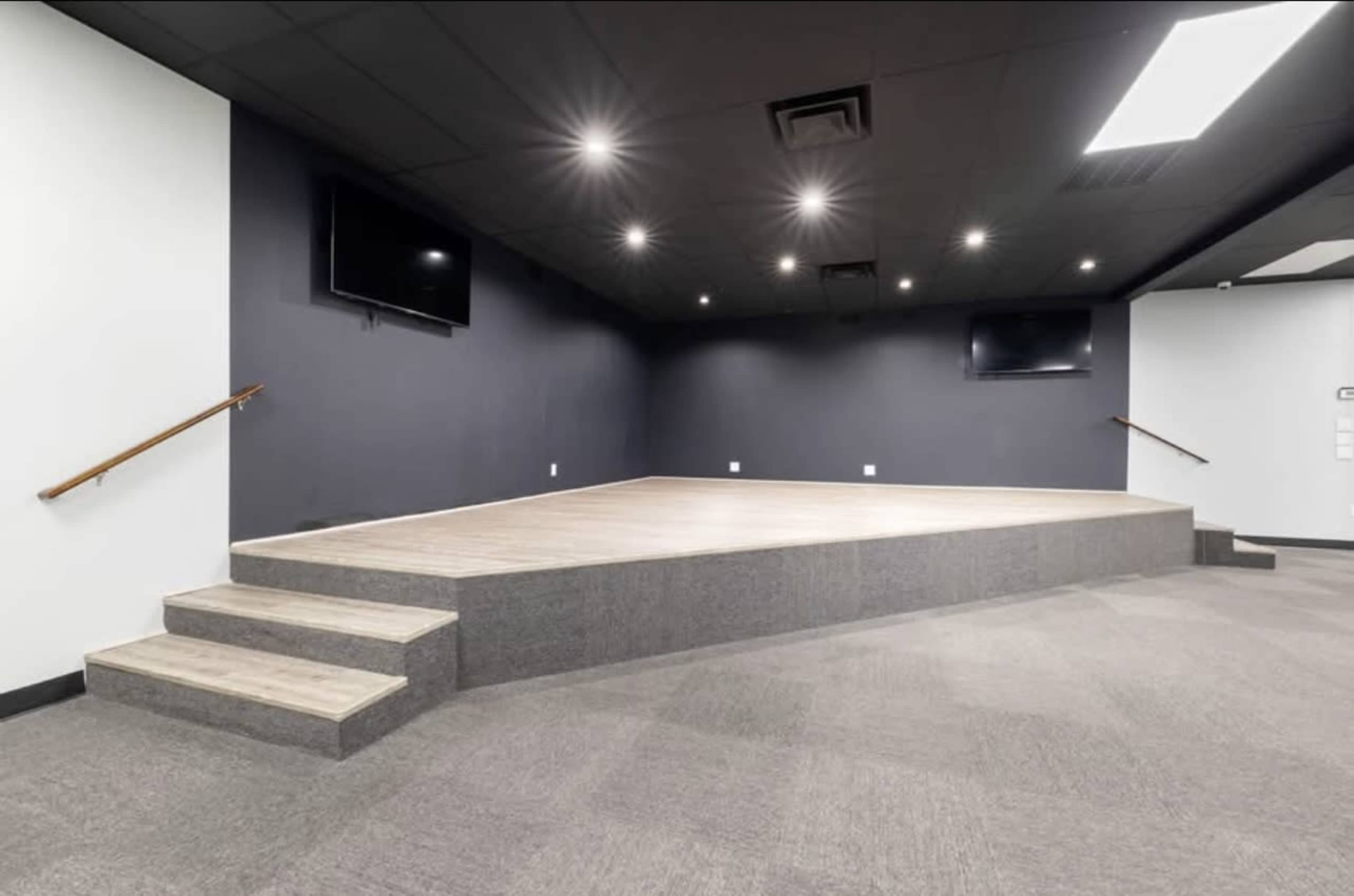 The image shows a spacious room with a raised stage, equipped with flat-screen TVs and a set of steps leading up to the stage area, all under a dark ceiling with recessed lighting.