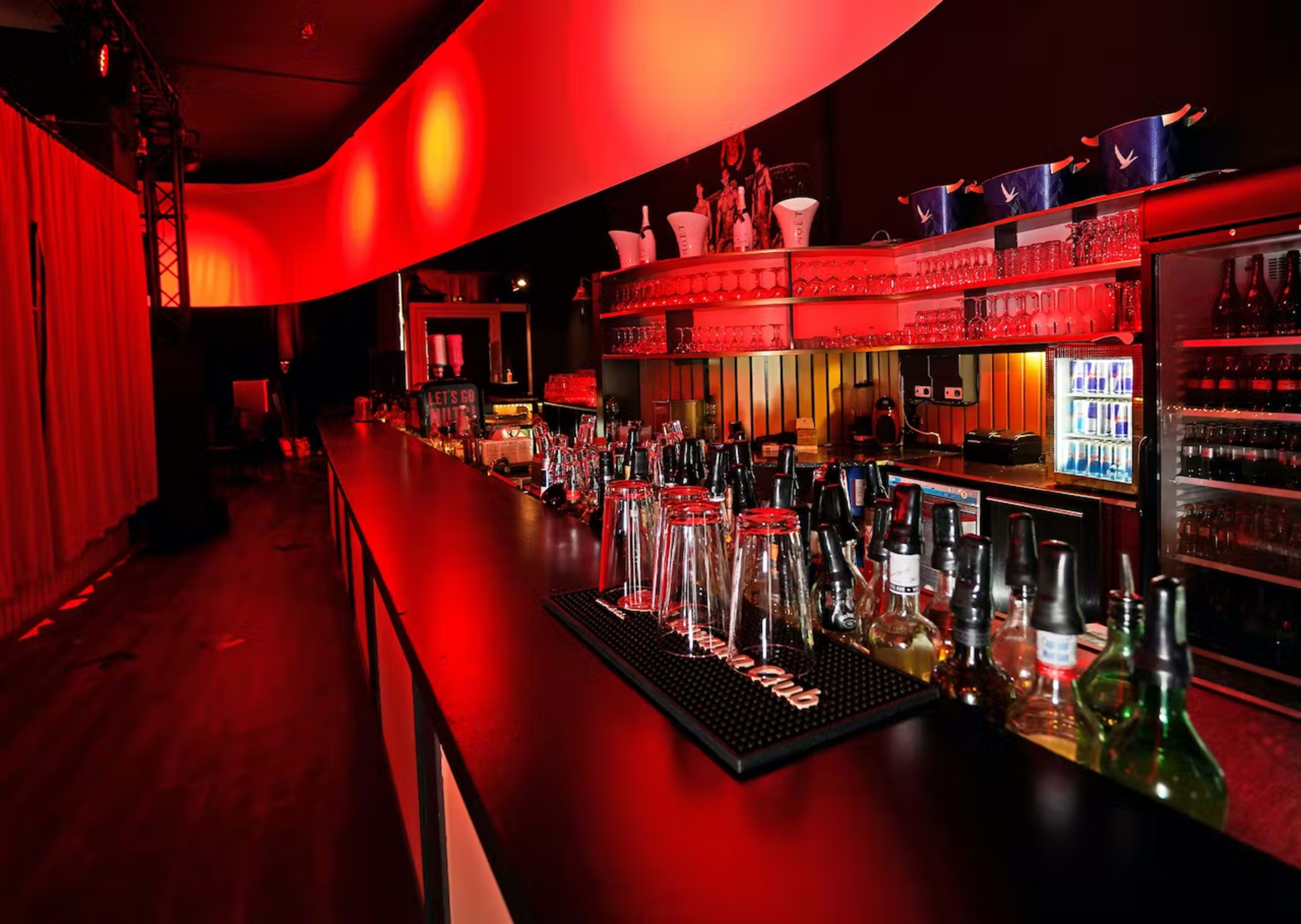 The image shows a modern bar with a long counter, illuminated by red lights, displaying various bottles and glassware on the shelves.