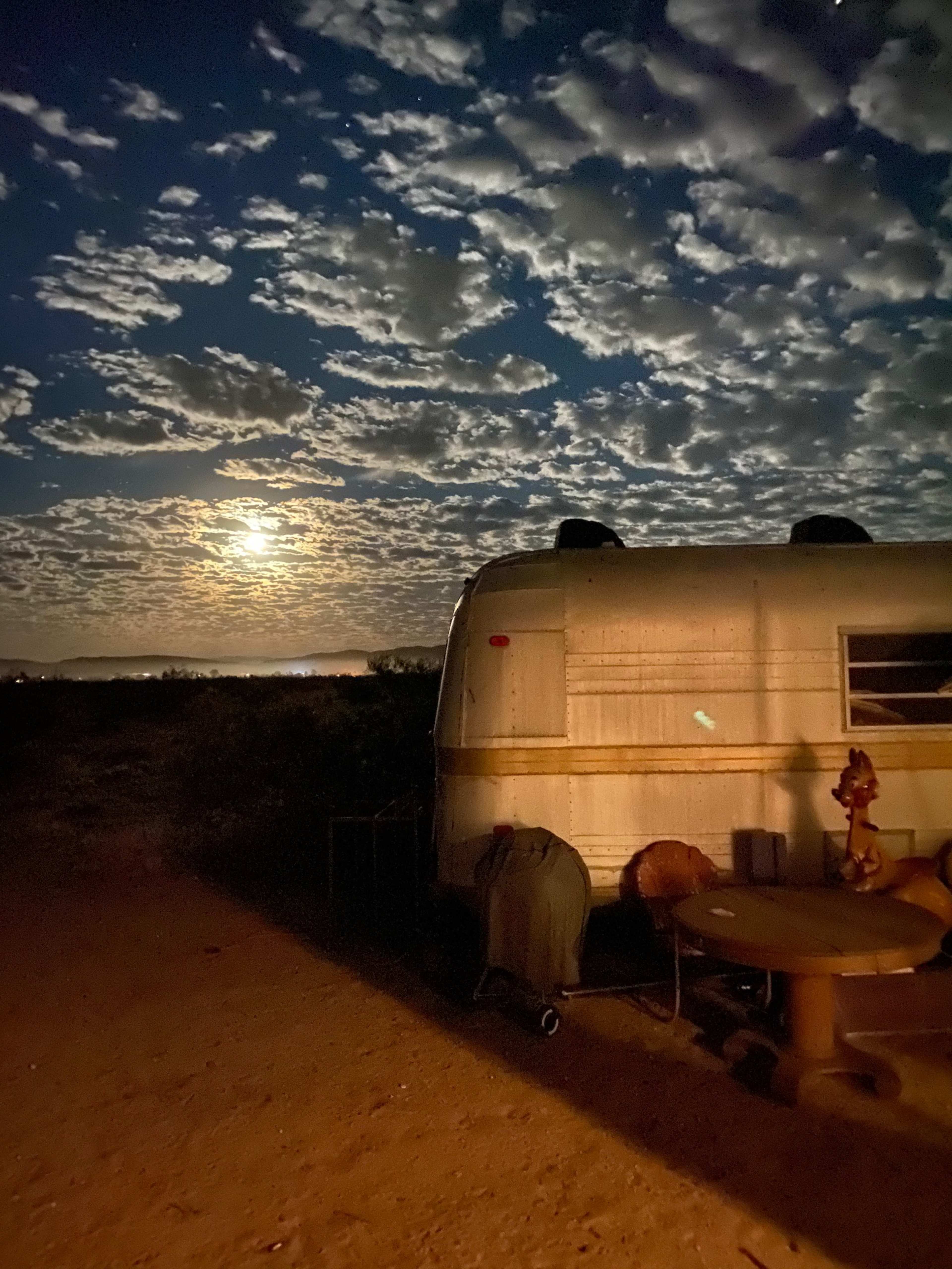 A vintage trailer is parked in a desert setting under a moonlit sky filled with clouds.