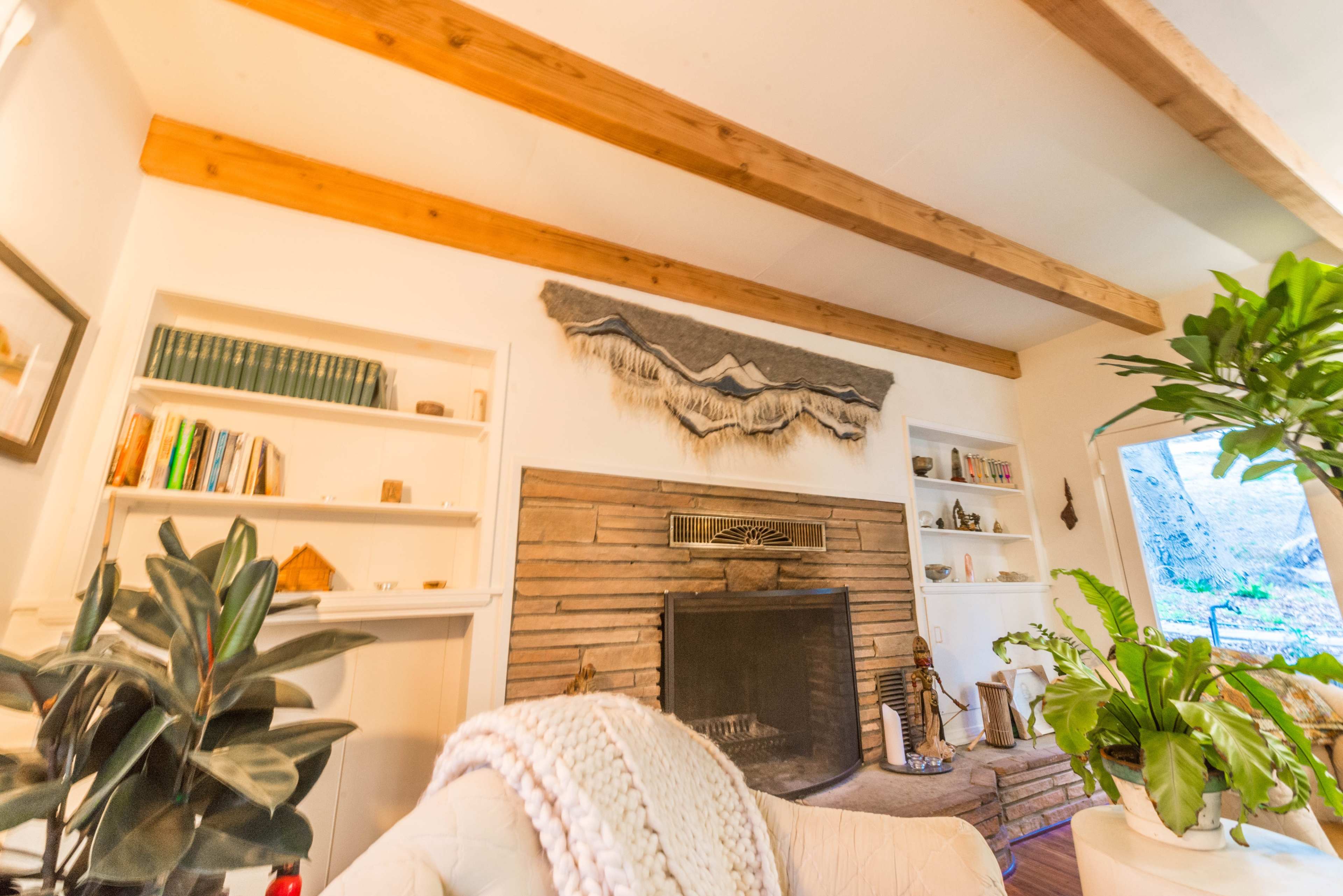 A living room with a stone fireplace, wooden beams on the ceiling, and bookshelves filled with books and decorative items, alongside indoor plants.
