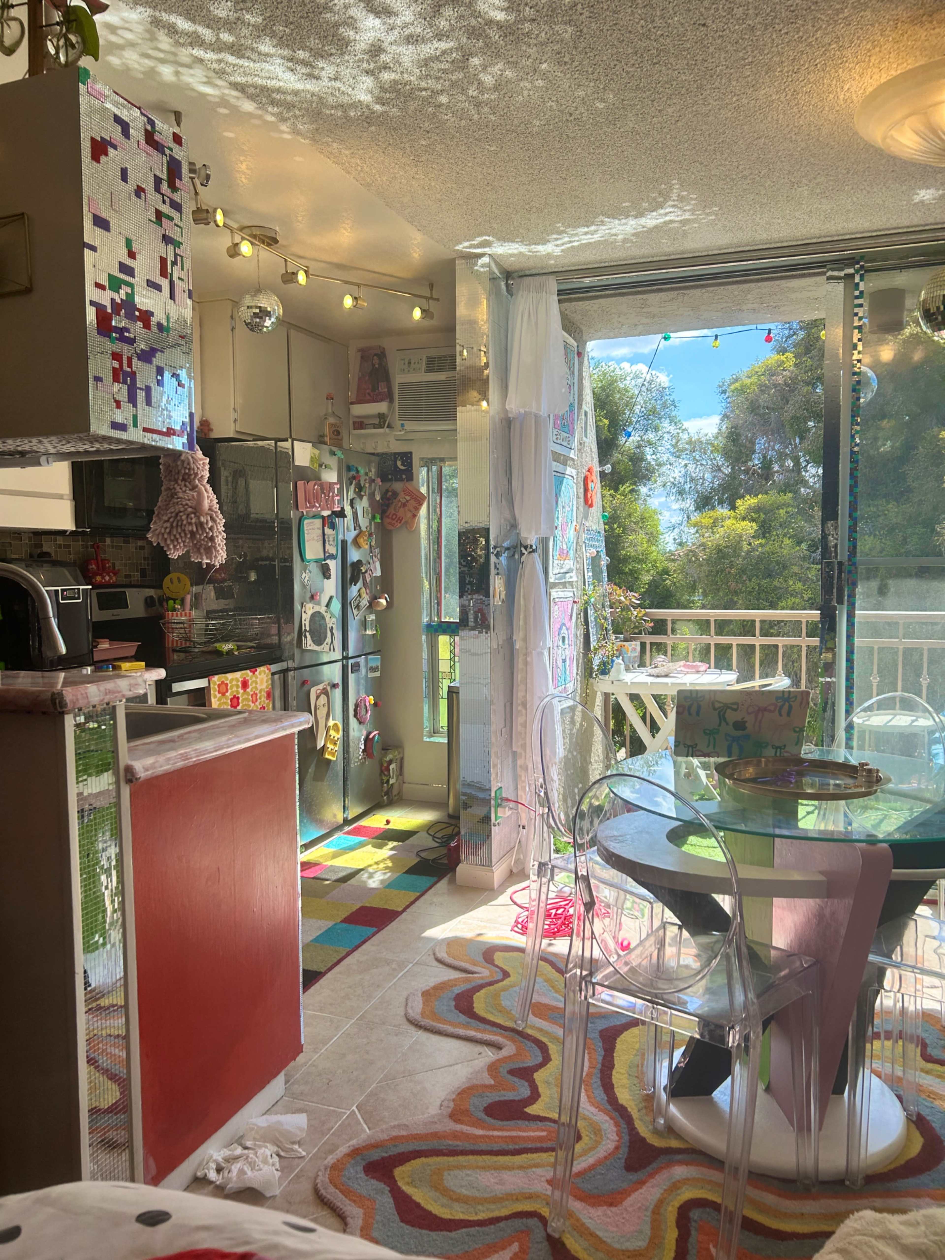 The image shows a brightly colored apartment interior with a kitchen area, a dining table with transparent chairs, and large windows letting in natural light.