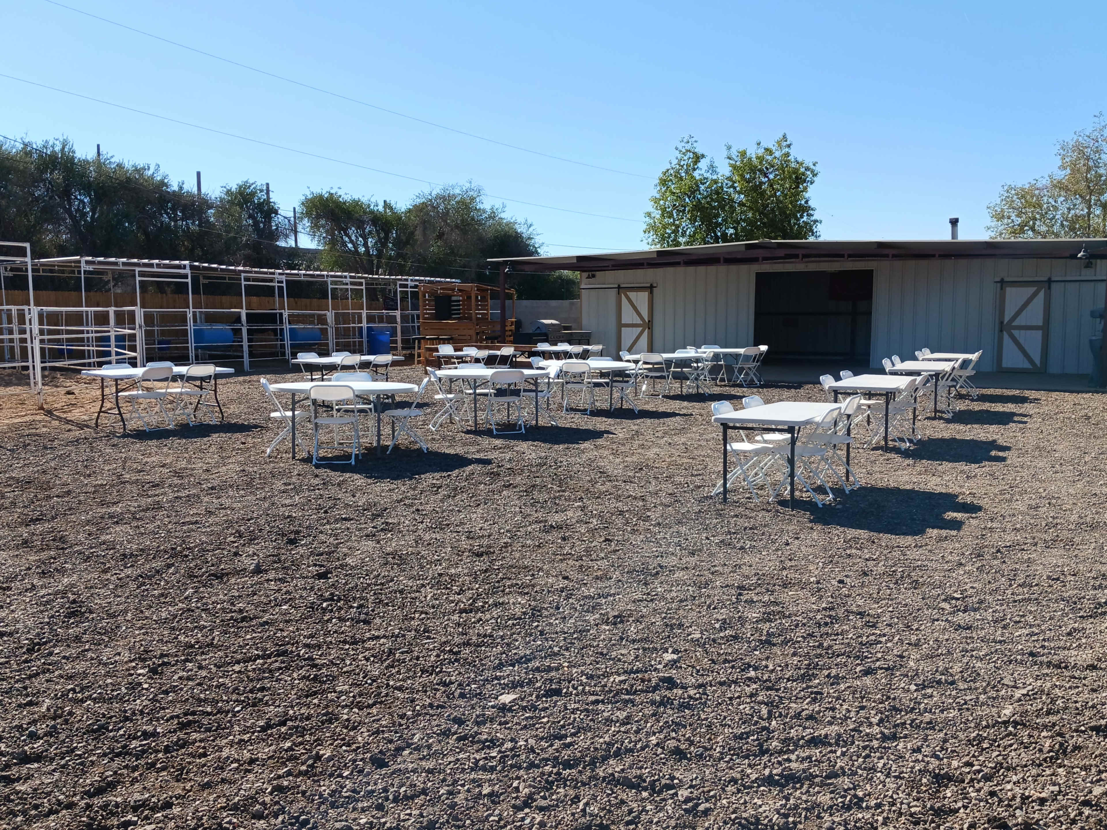 A spacious outdoor area with multiple folding tables arranged on a gravel surface, adjacent to a building with open doors.