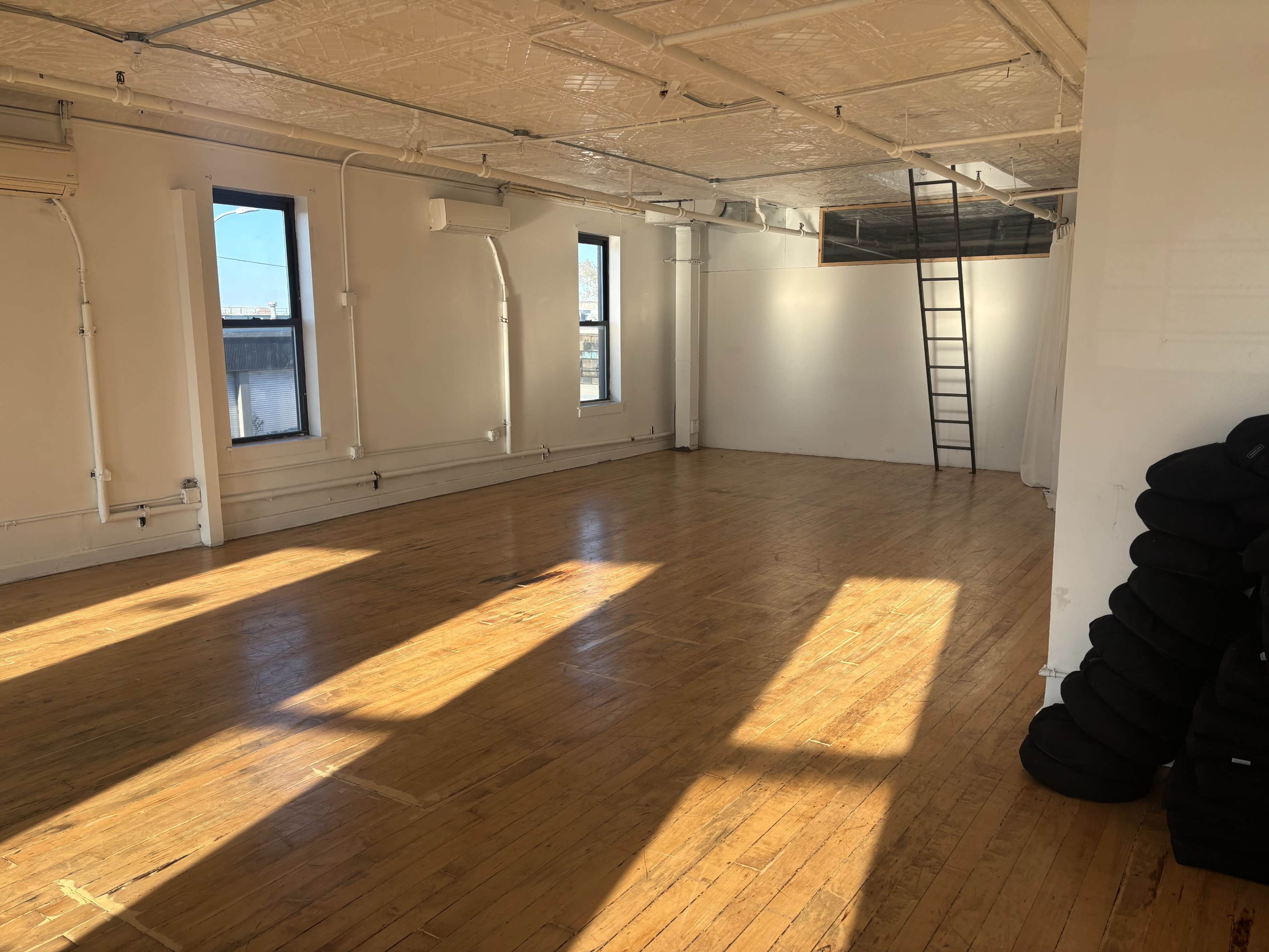The image shows a spacious, empty room with wooden floors, two windows letting in natural light, and a ladder leading to a loft area.