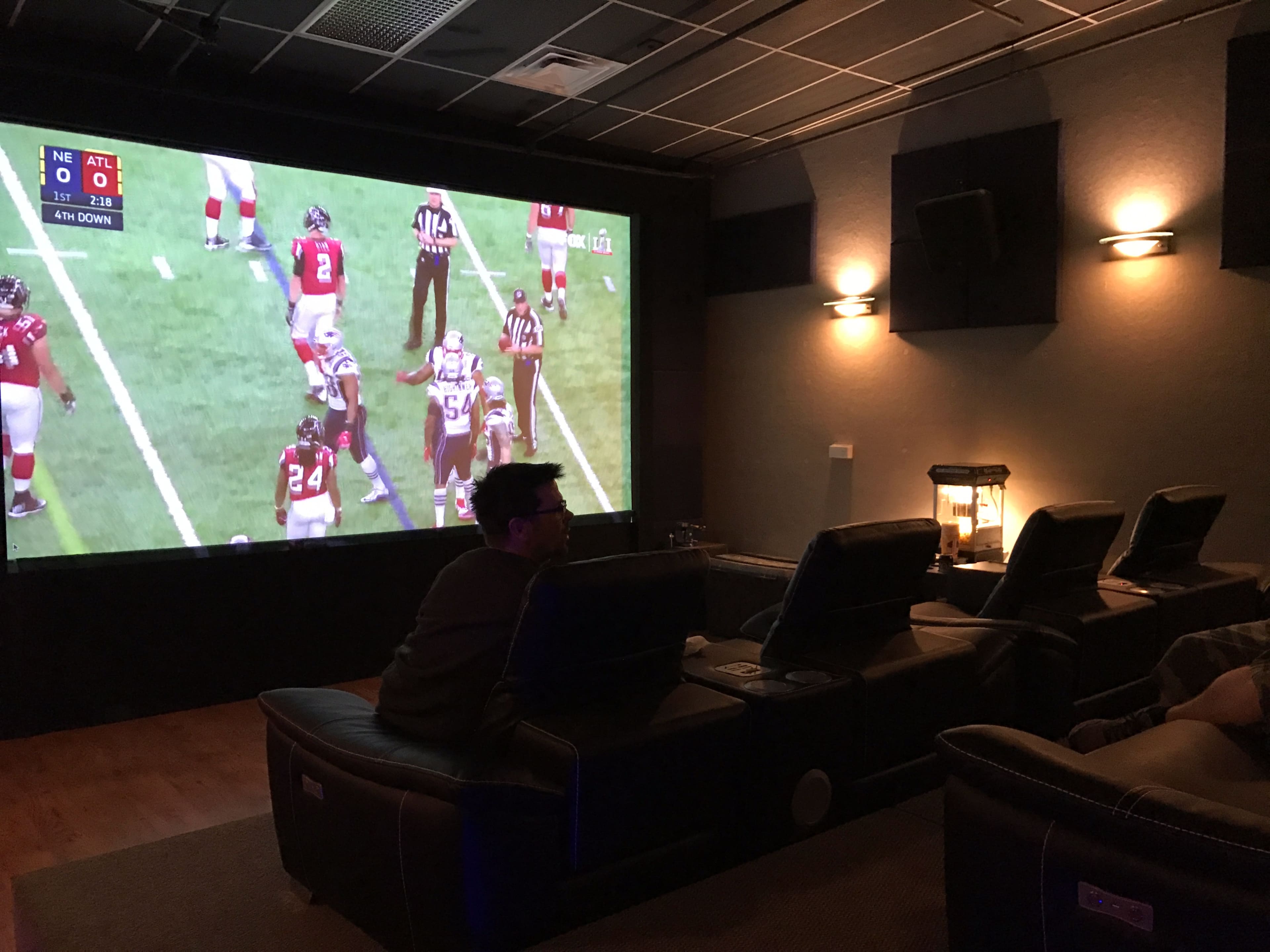 A group of people watches a football game on a large screen in a home theater with reclining seats and ambient lighting.