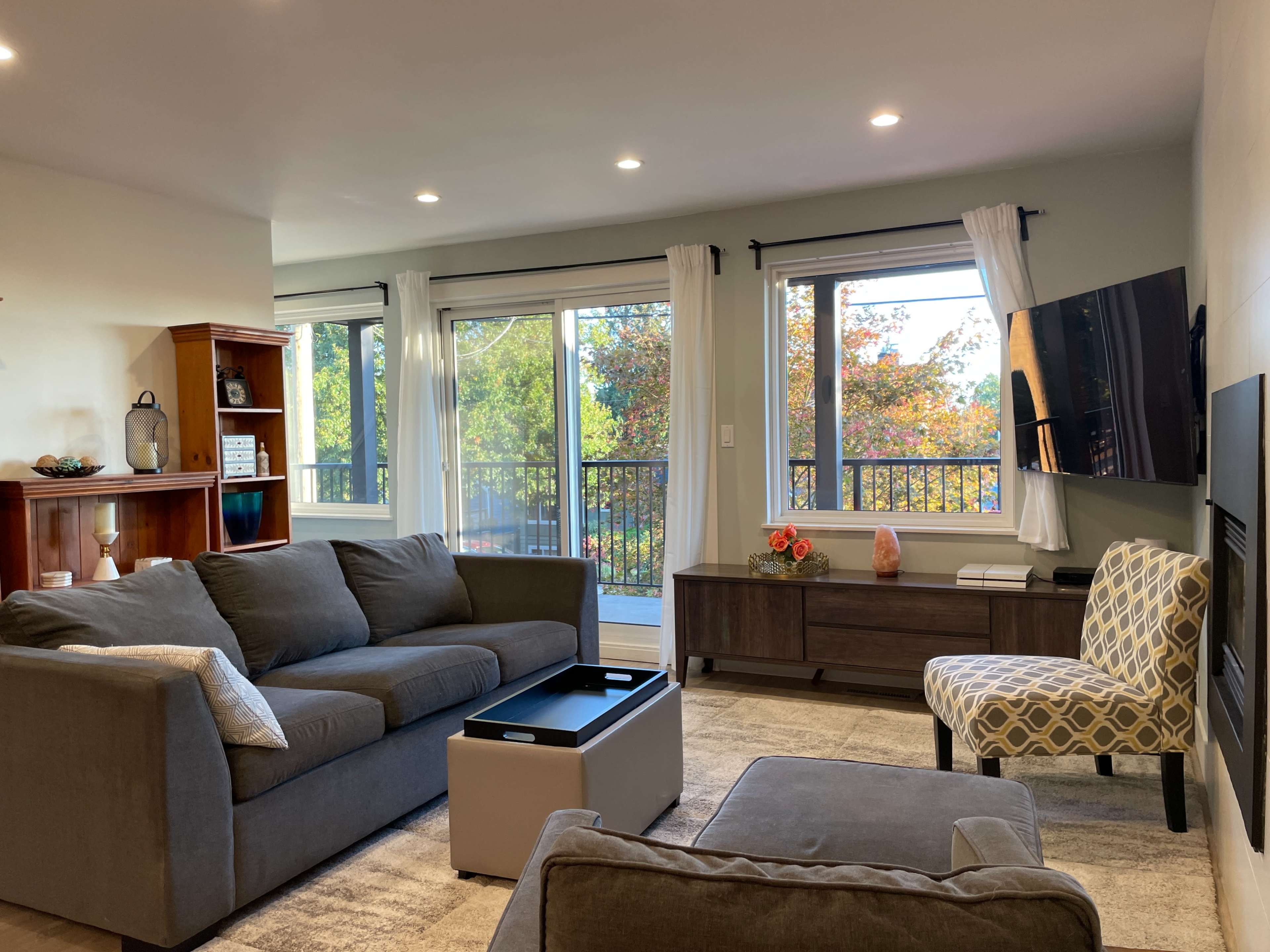 The living room features two gray couches, a coffee table, a mounted TV, and large windows overlooking a tree-filled view.