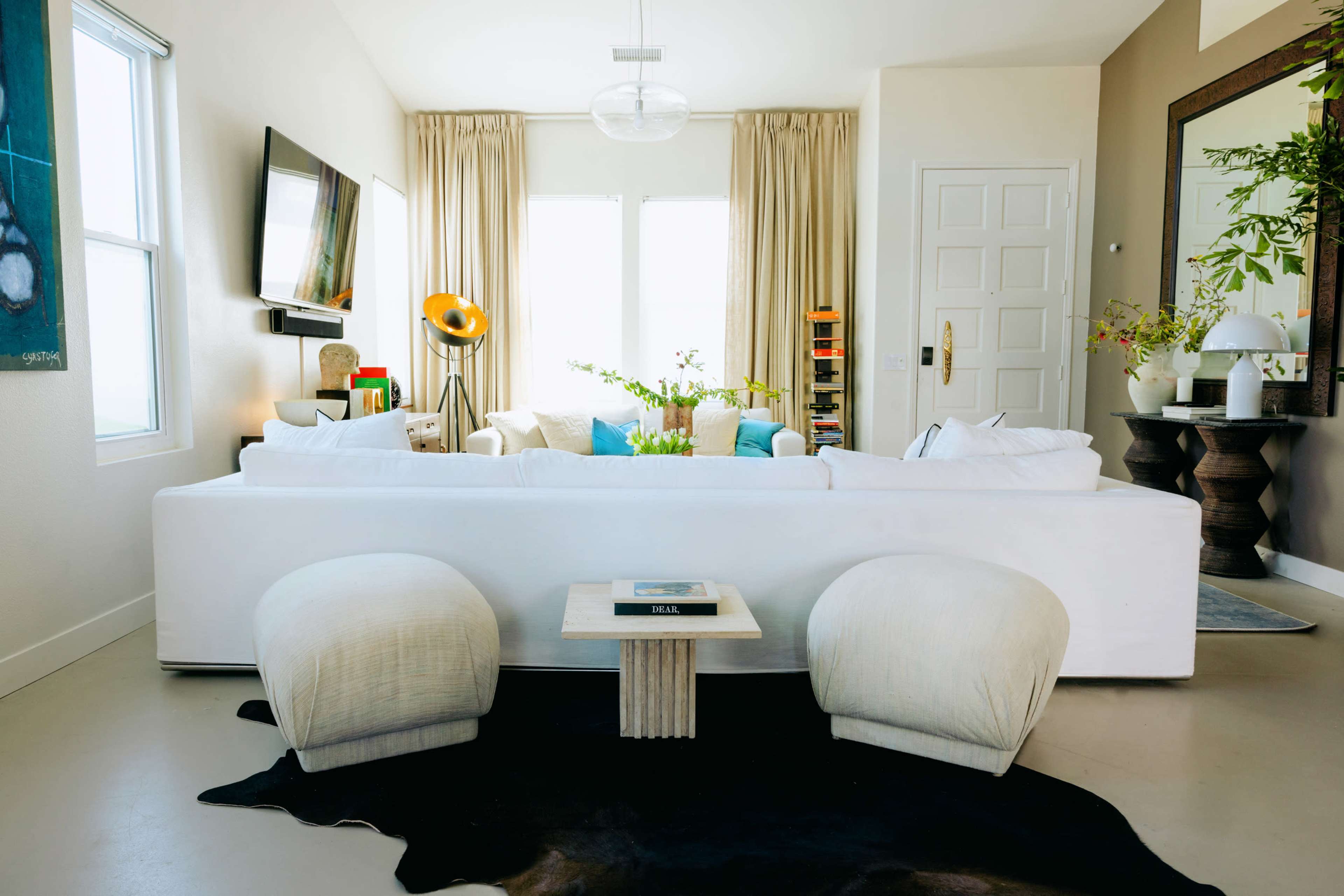 A modern living room with a white sofa, two round upholstered chairs, a wooden coffee table, and large windows covered with light curtains.