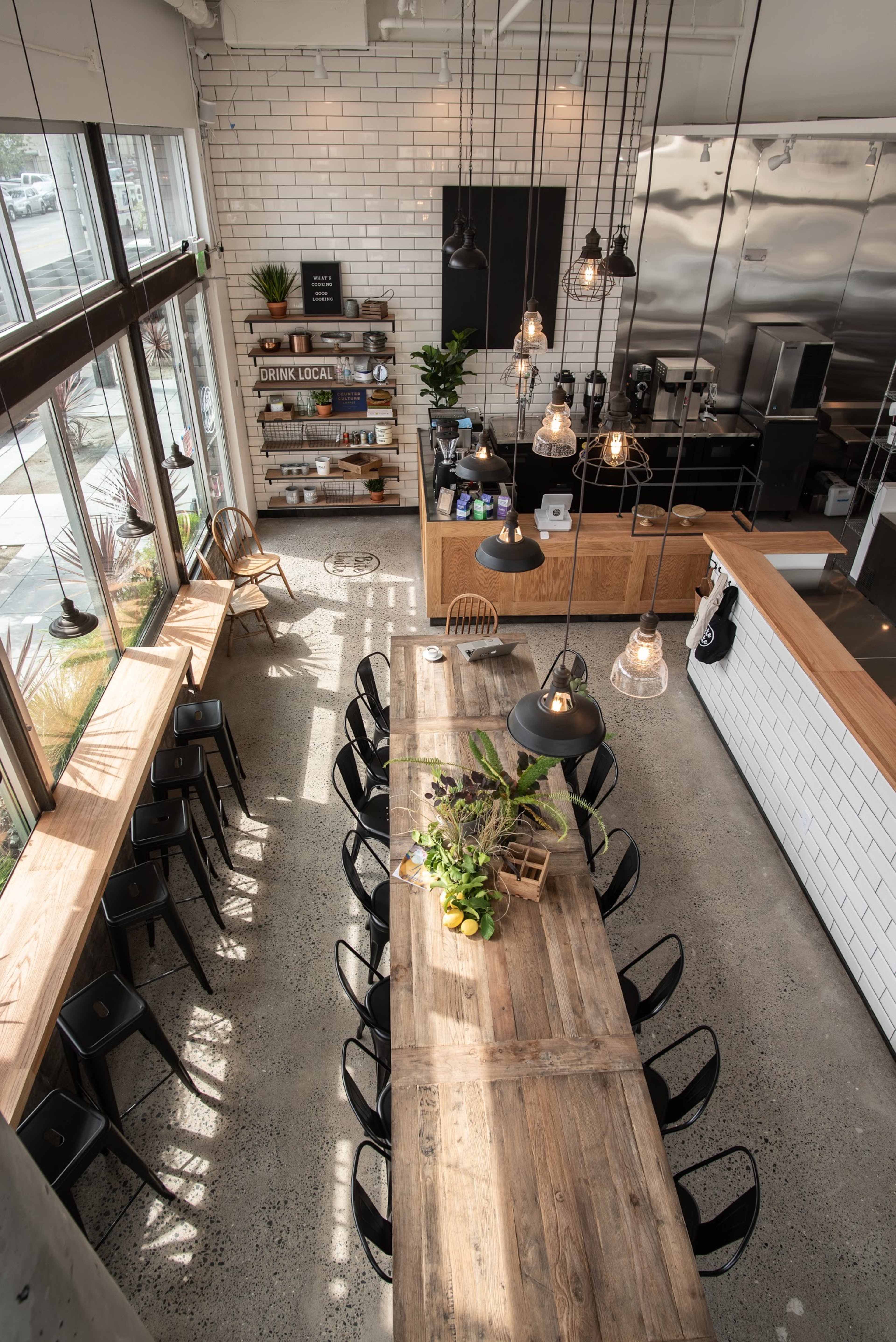 An industrial-style cafe with a long wooden table, black chairs, large windows, and pendant lighting.