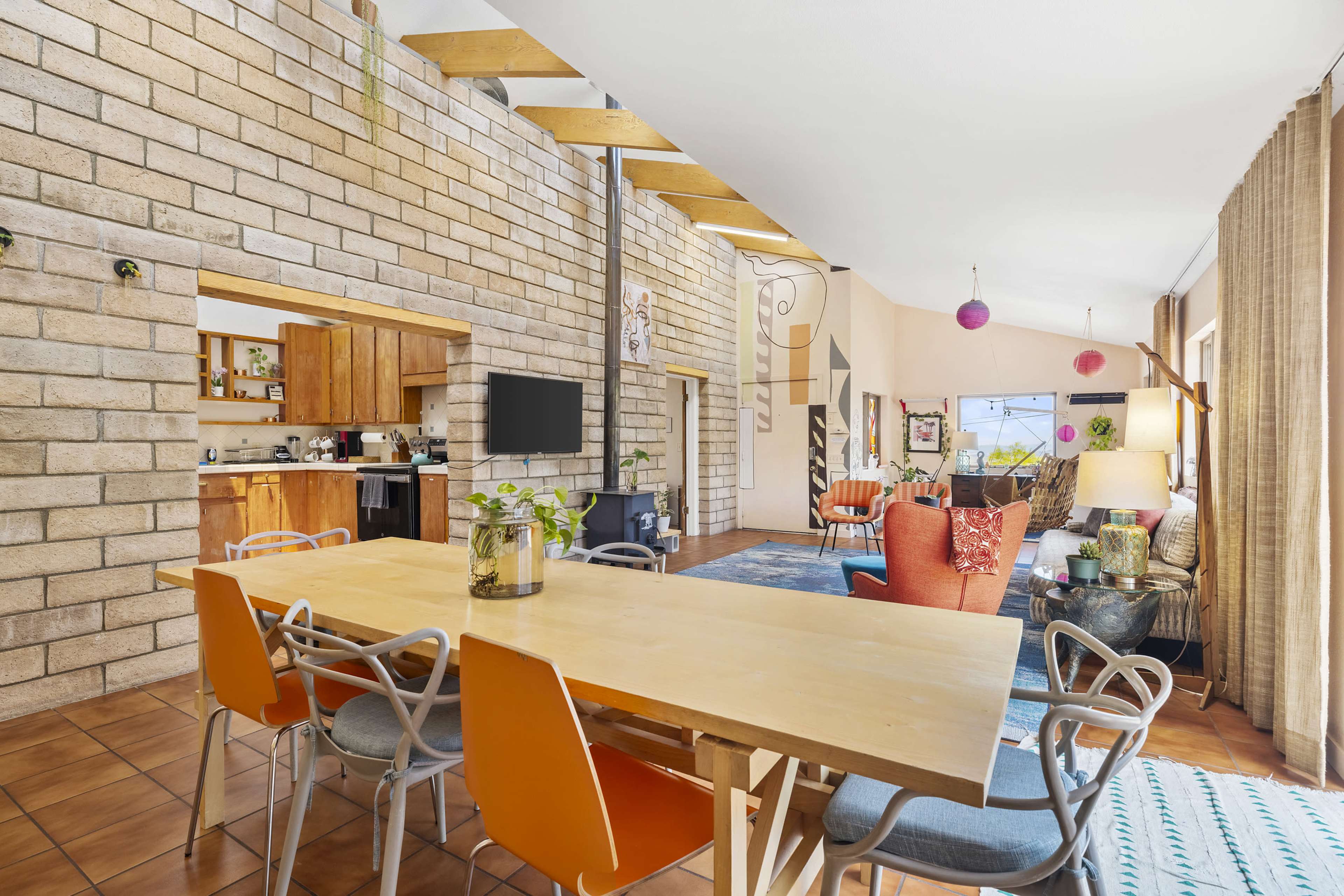 The image shows a modern interior space featuring a dining table with orange chairs, an open kitchen, and a living area with colorful decor and large windows.