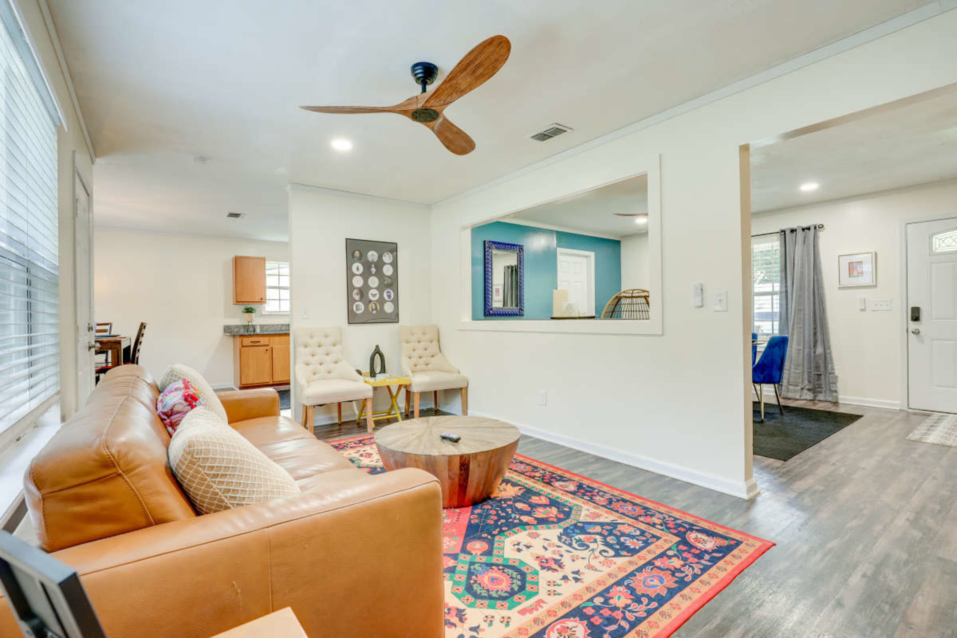 A light-filled living room features a brown leather sofa, a round coffee table, and a colorful area rug, with a ceiling fan and a window to the kitchen.
