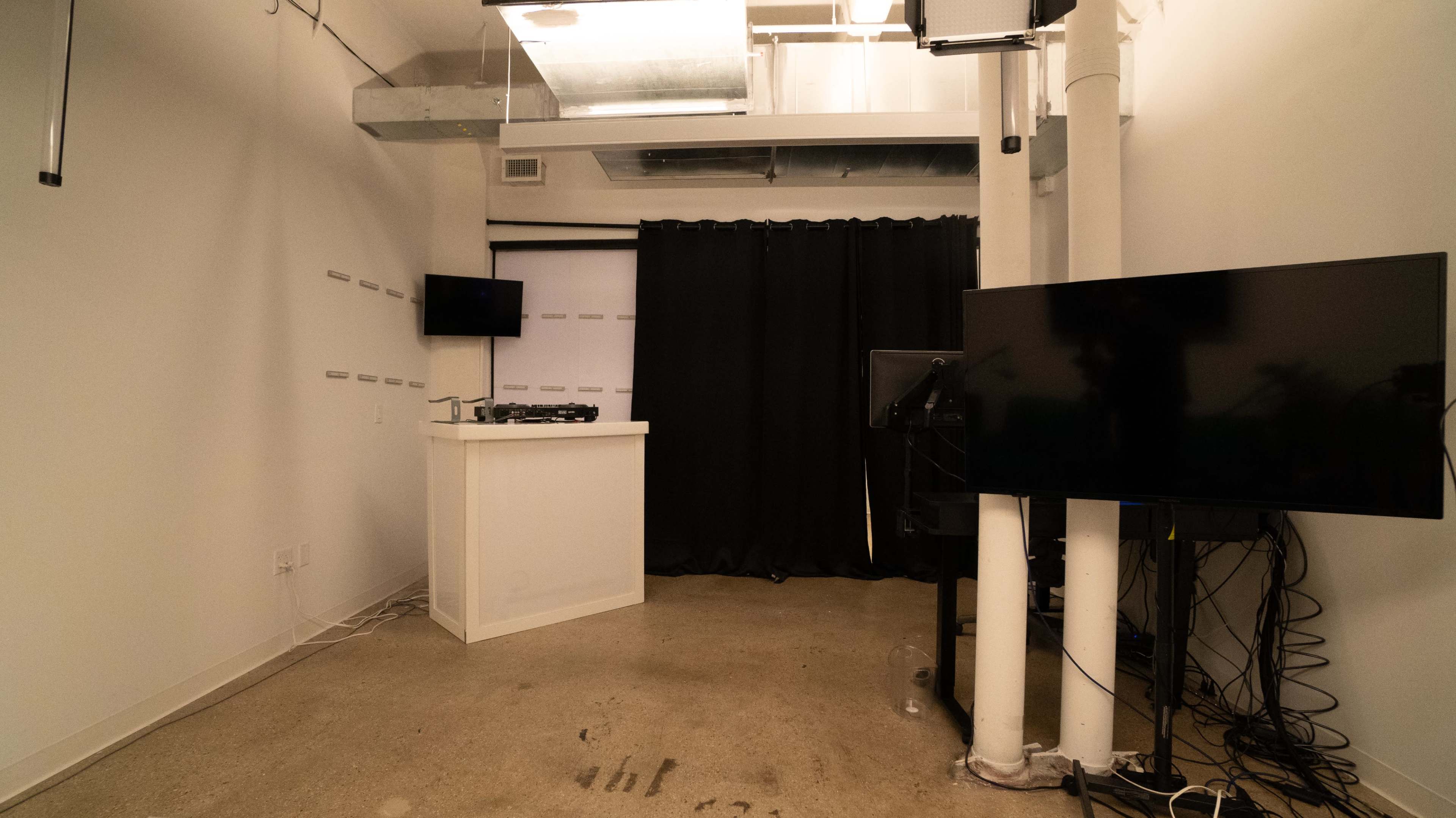 The image shows a minimalist room with a white counter, two mounted televisions, and a black curtain dividing the space.
