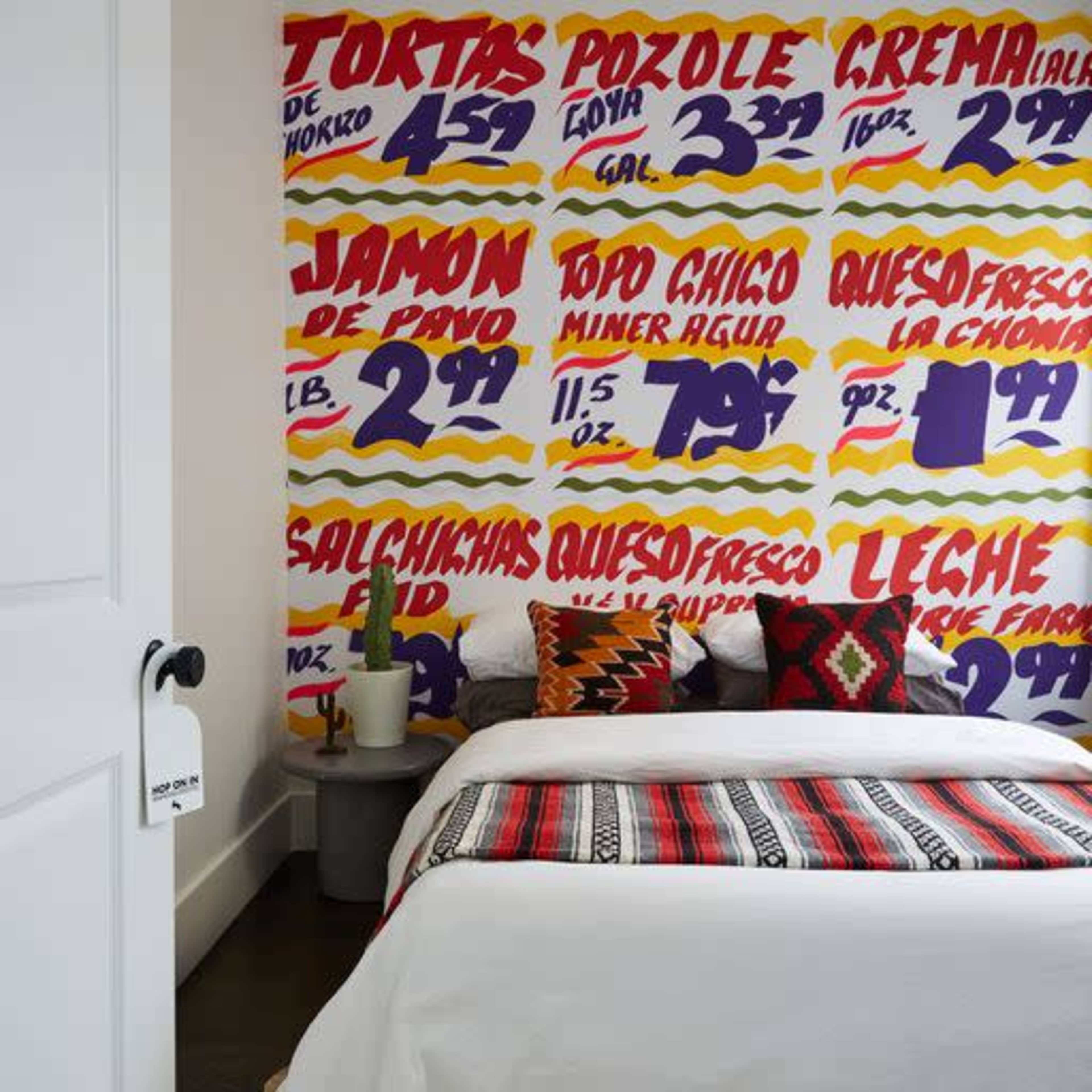The image depicts a bedroom with a brightly colored mural of food items on the wall, a bed with a patterned blanket, and a small table with a potted plant.