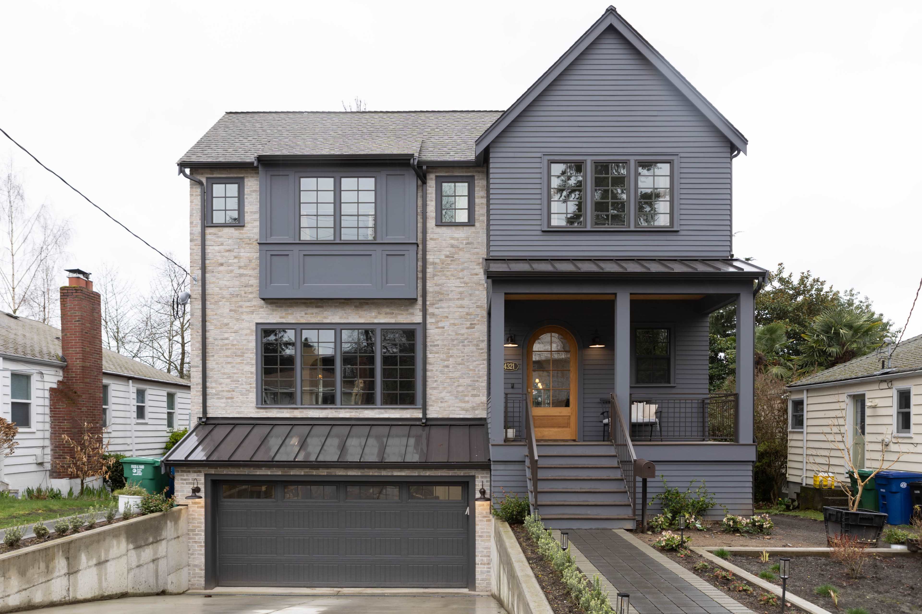 The image shows a two-story house with gray siding and brick accents, featuring a garage beneath the main structure and a porch with steps leading to the front door.