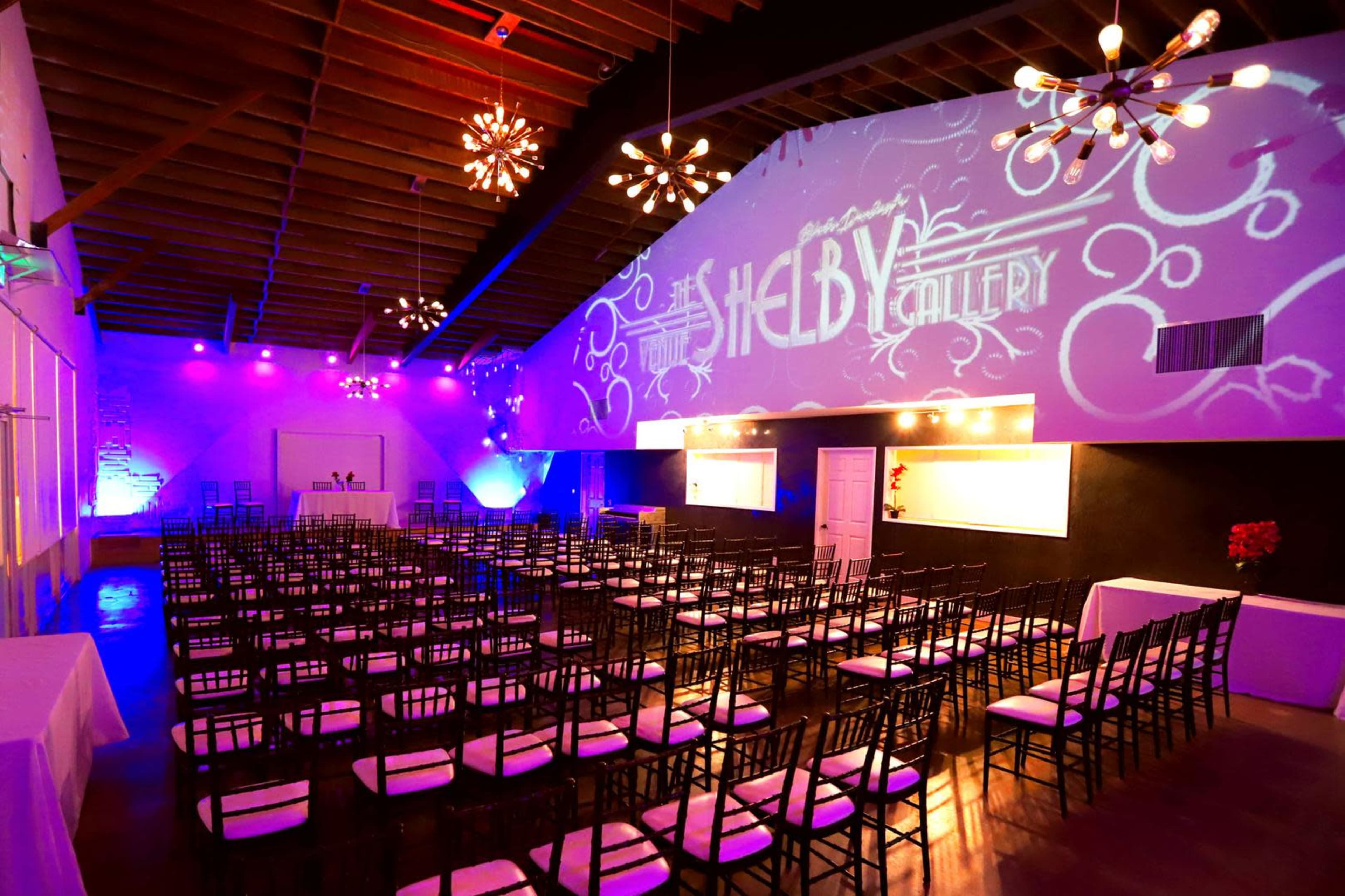The image shows a banquet hall set up with rows of black chairs facing a stage, illuminated by colorful lighting and featuring a projected logo on the back wall.