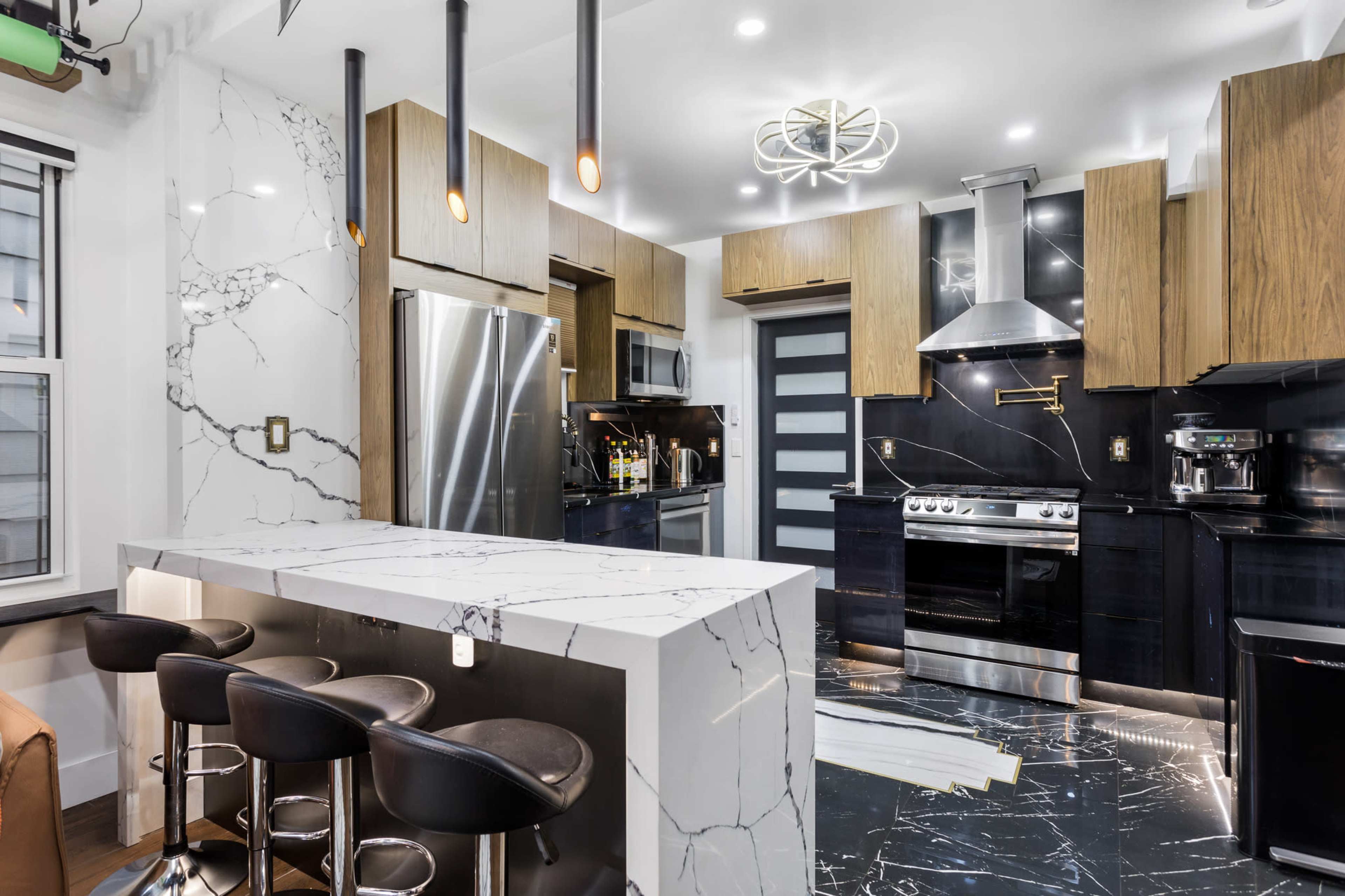 A modern kitchen featuring marble countertops, wooden cabinetry, and stainless steel appliances, along with a bar area with black stools.