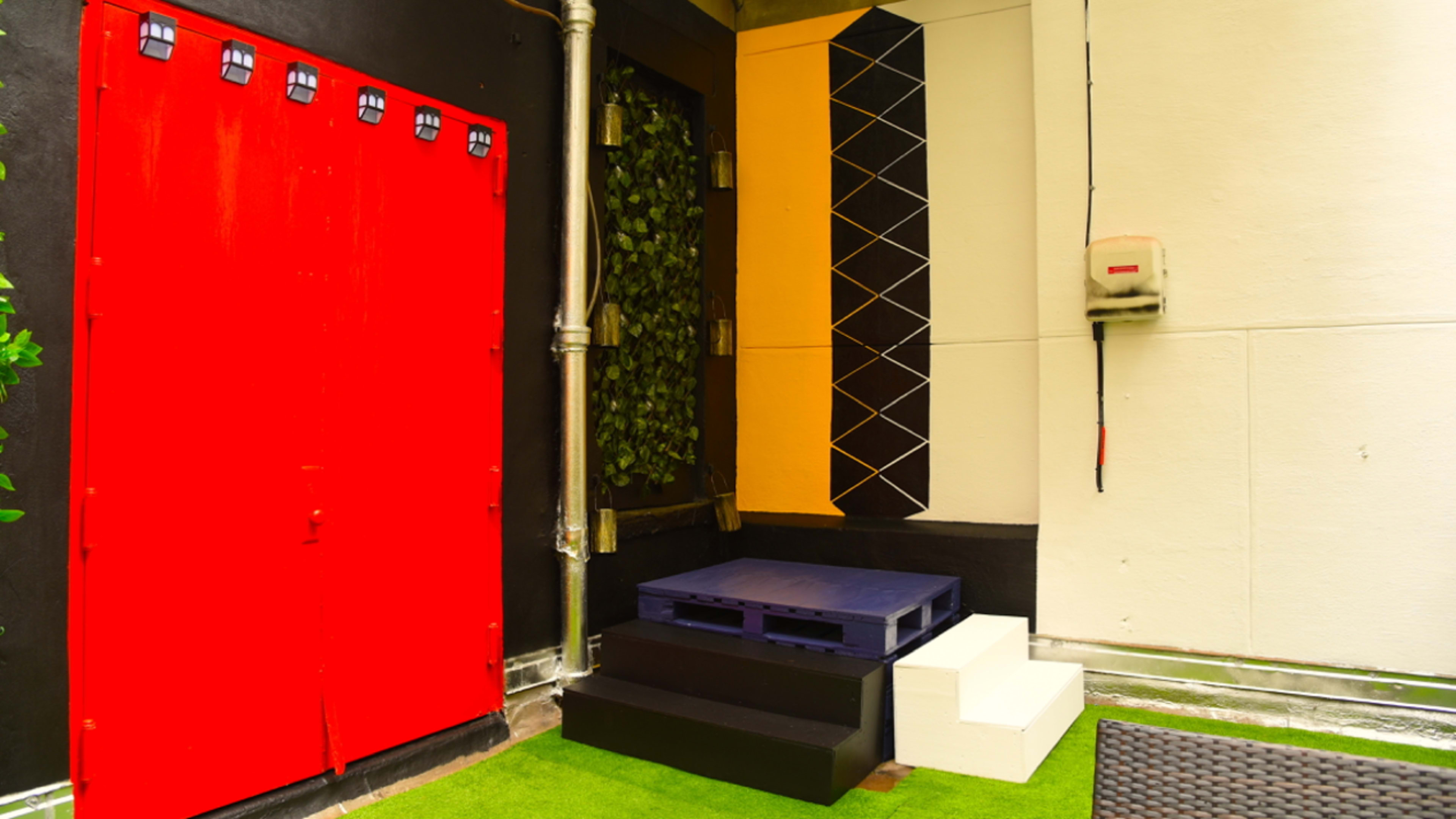 A small outdoor space with a red door, a black wall adorned with greenery, and steps leading up to a platform made of pallets and painted white.