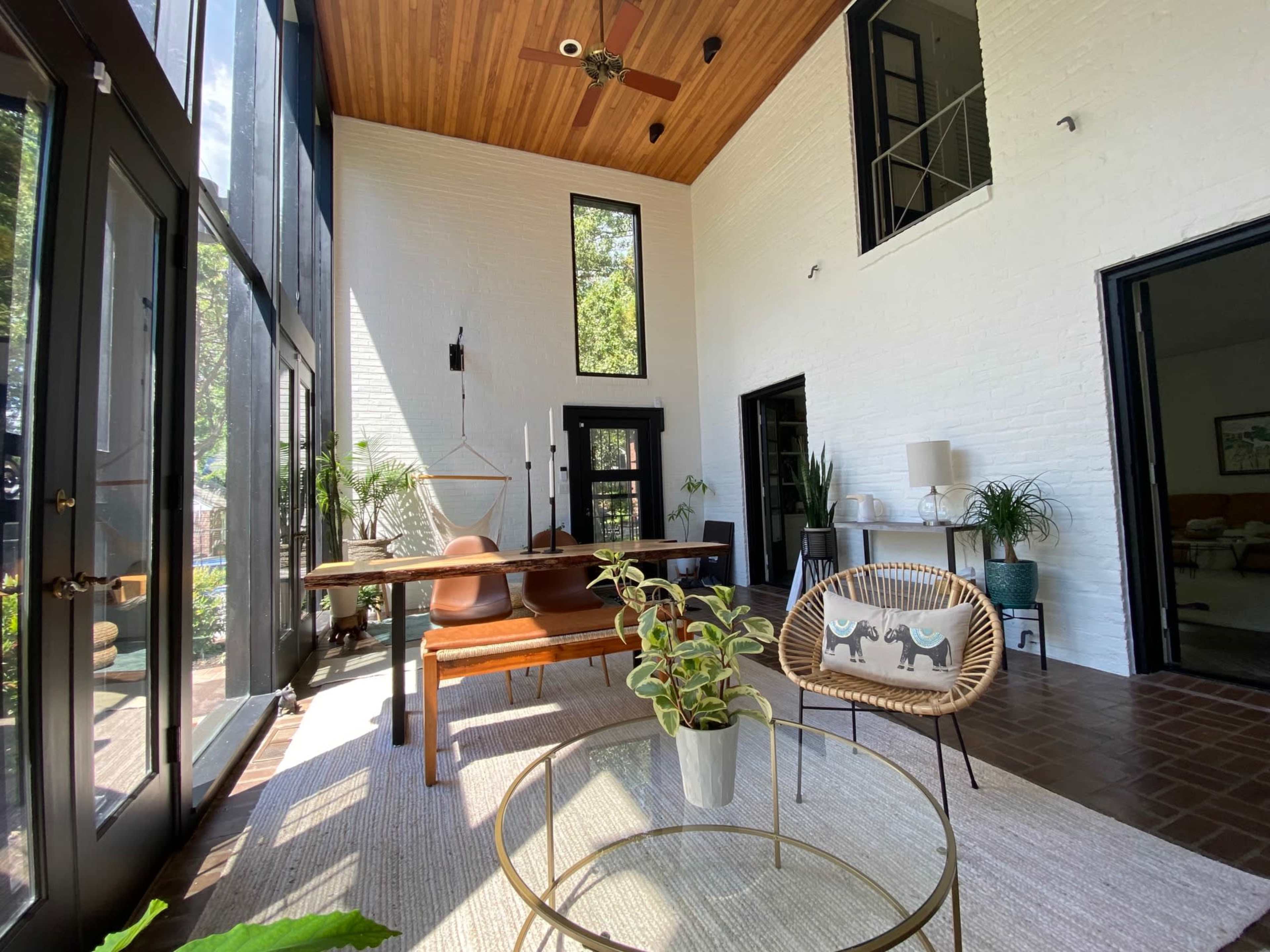 A bright, open living space features a dining table, chairs, and plants, with large windows allowing ample natural light.