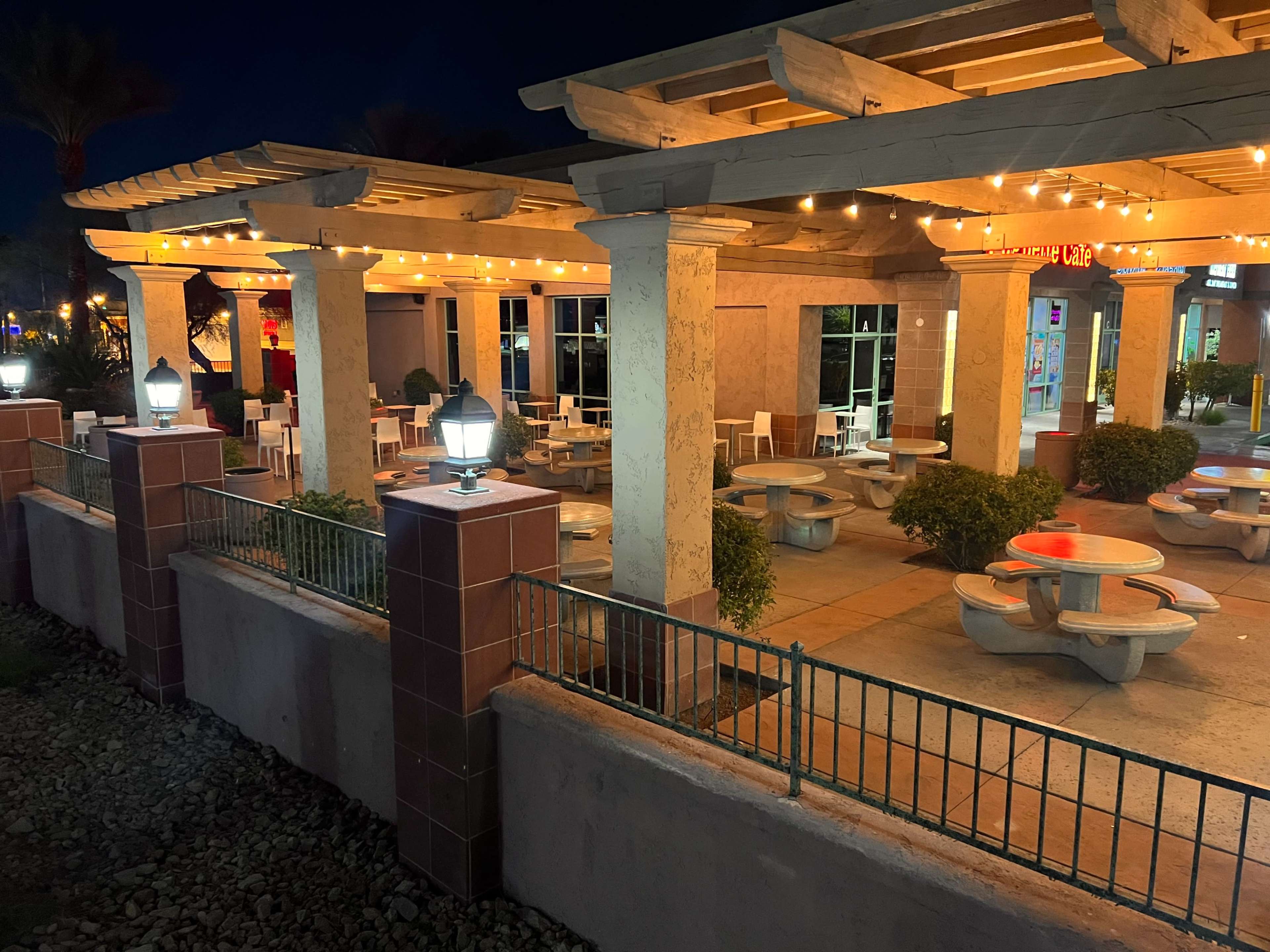 A partially illuminated outdoor dining area with several table and chair setups under a covered structure, framed by decorative lighting.