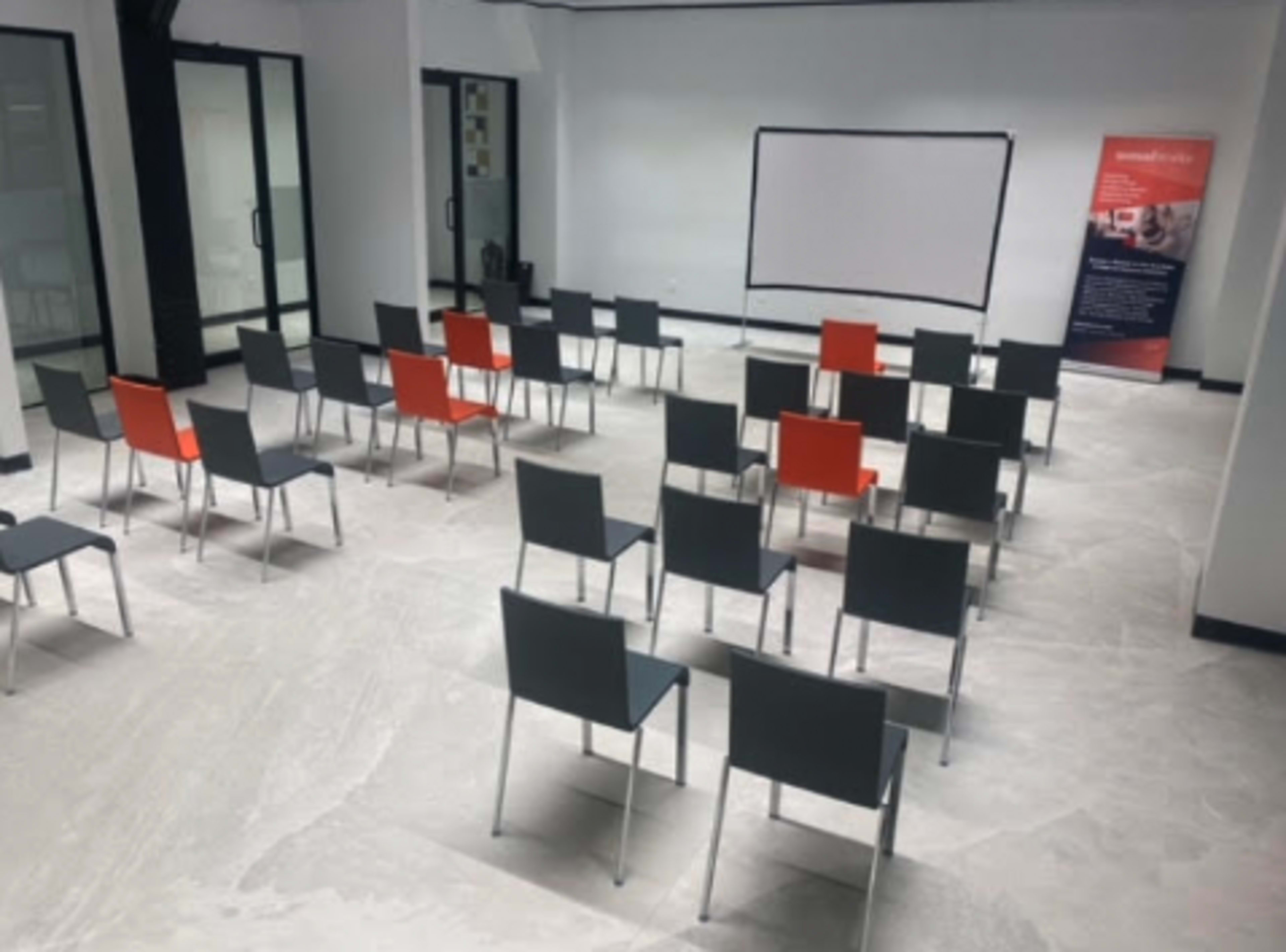 A training room is arranged with rows of gray and orange chairs facing a white screen and a banner in the background.