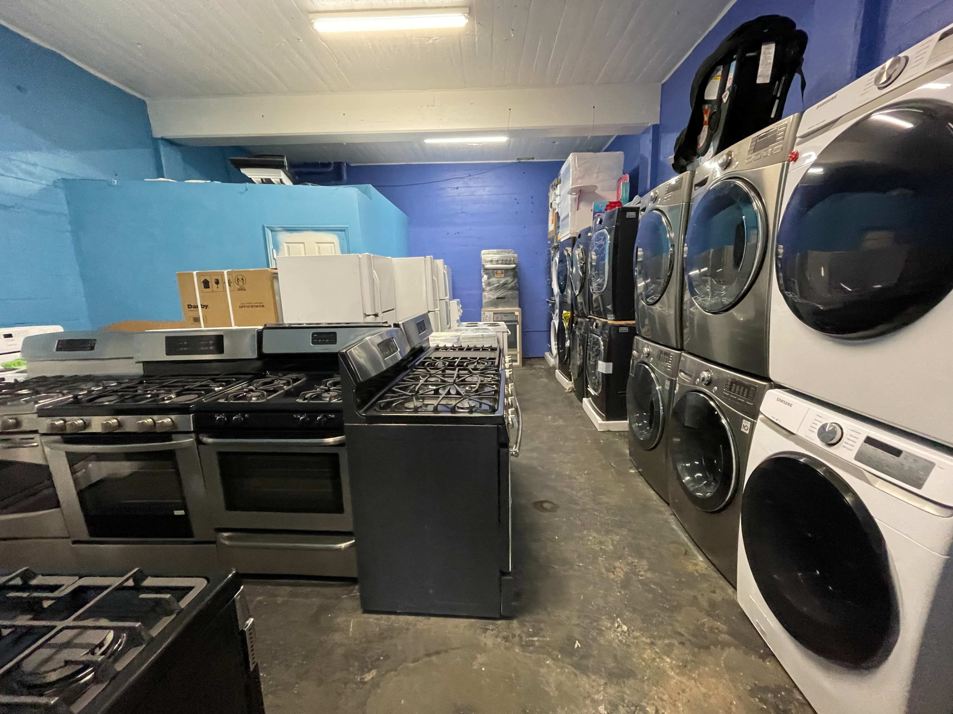 The image shows a showroom displaying various kitchen and laundry appliances, including stoves, refrigerators, and washing machines, against a blue wall.
