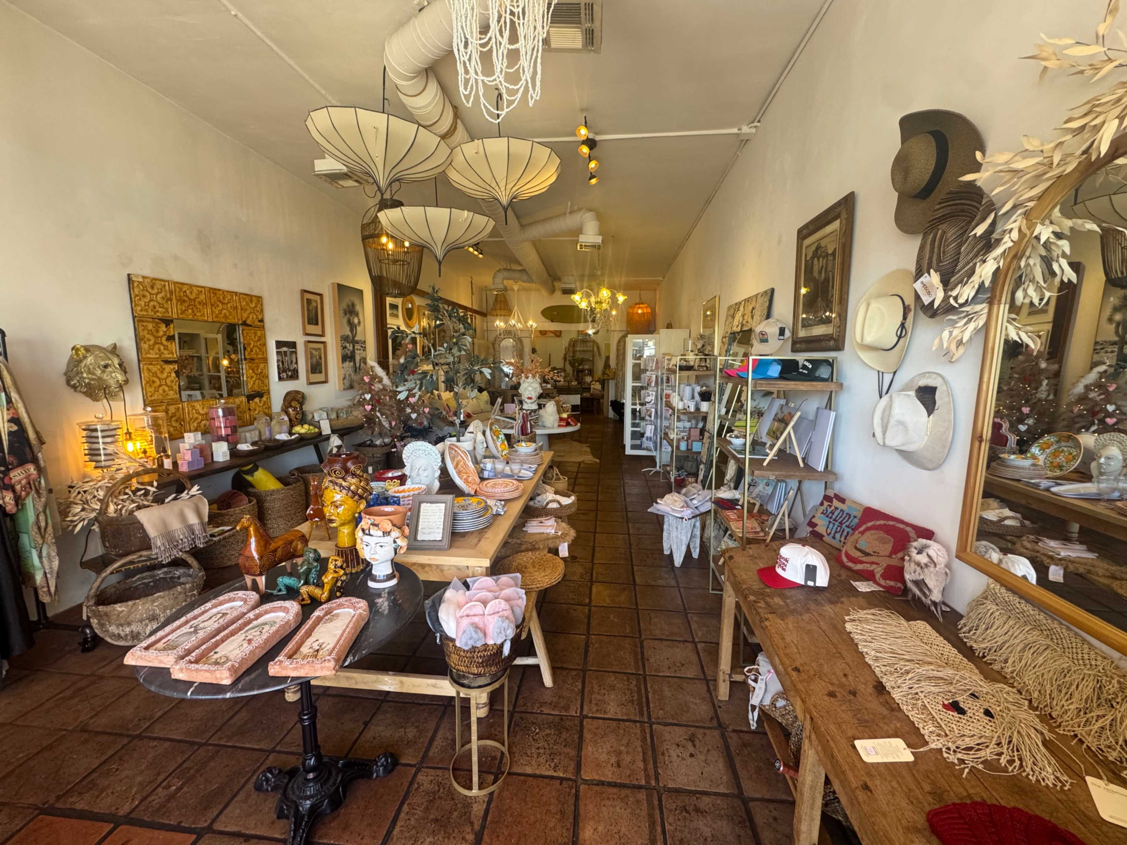 The image shows a cozy boutique filled with various decorative items, hats, and art pieces arranged on tables and shelves under ambient lighting.