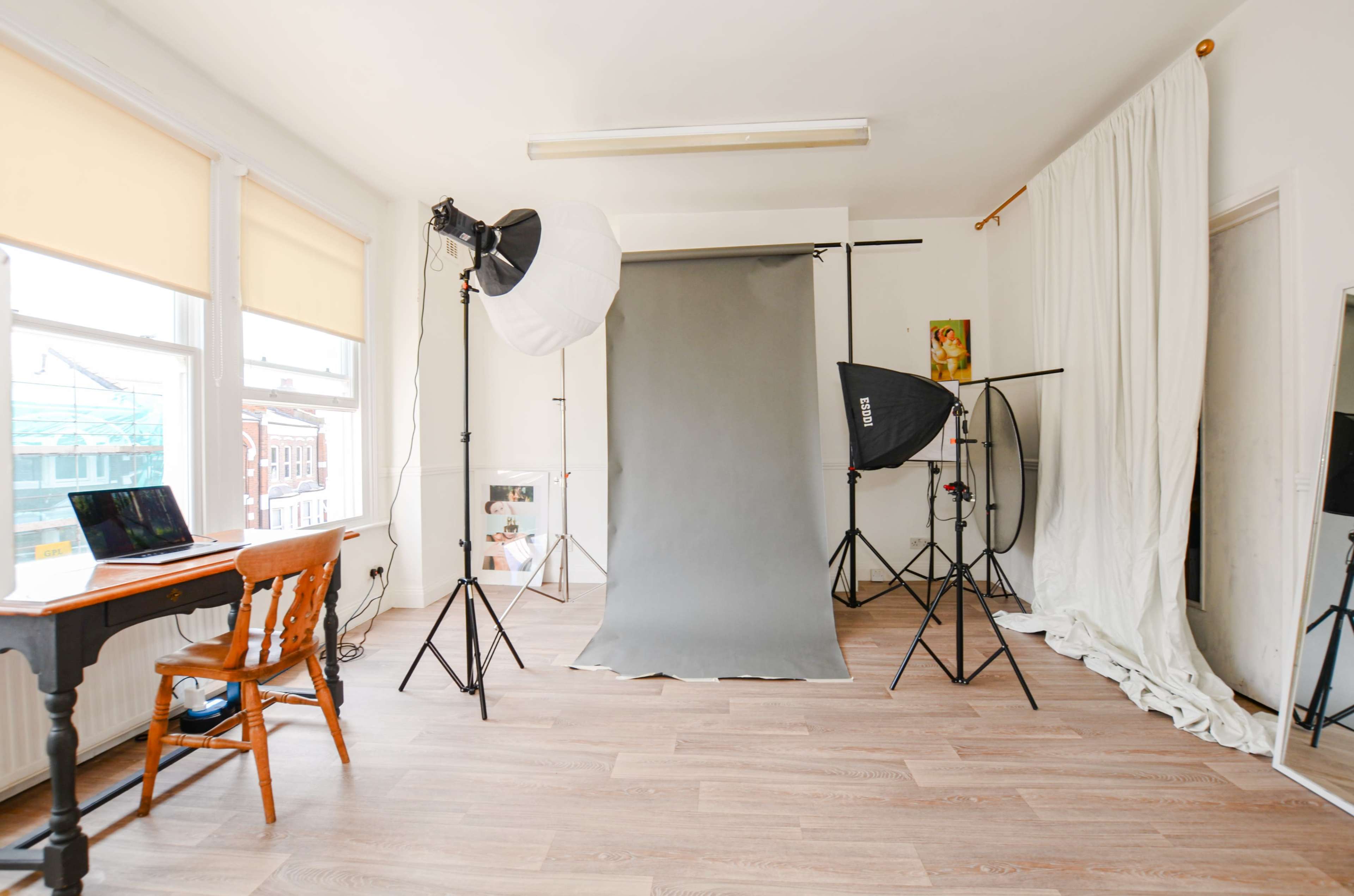 The image shows a photography studio equipped with a gray backdrop, lighting equipment, a wooden table with a laptop, and large windows allowing natural light to enter.