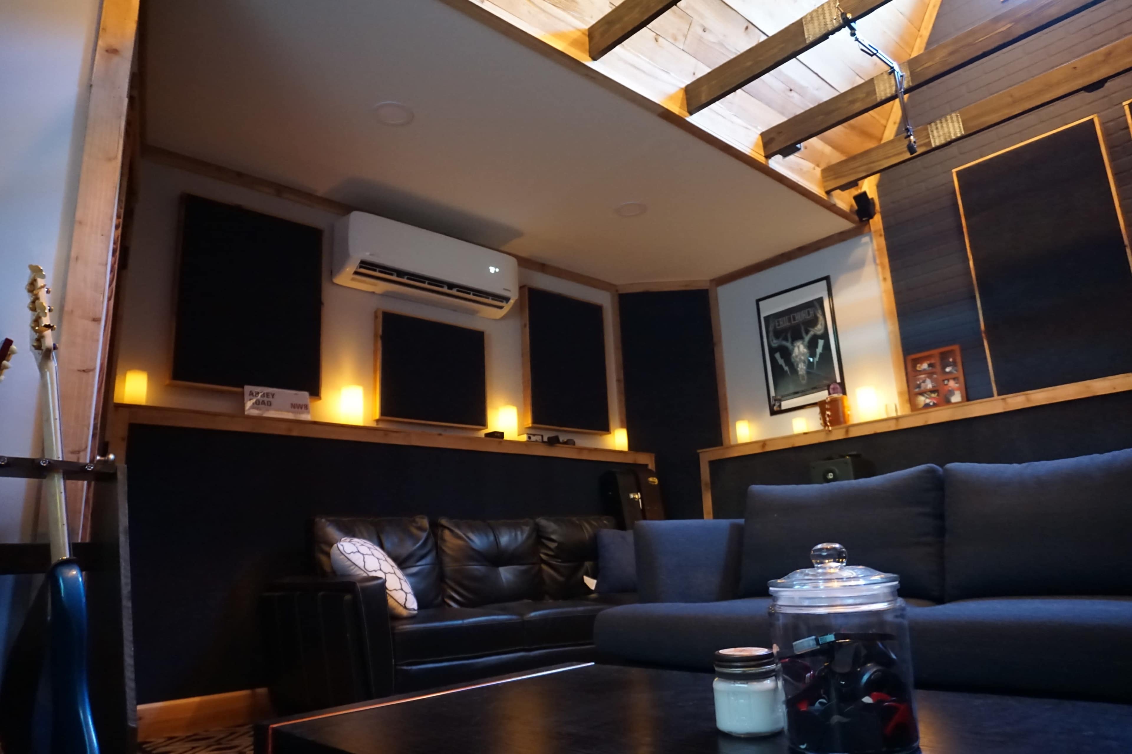 A cozy recording studio lounge featuring a black leather sofa, wooden beams, and soundproof panels, illuminated by soft lighting.