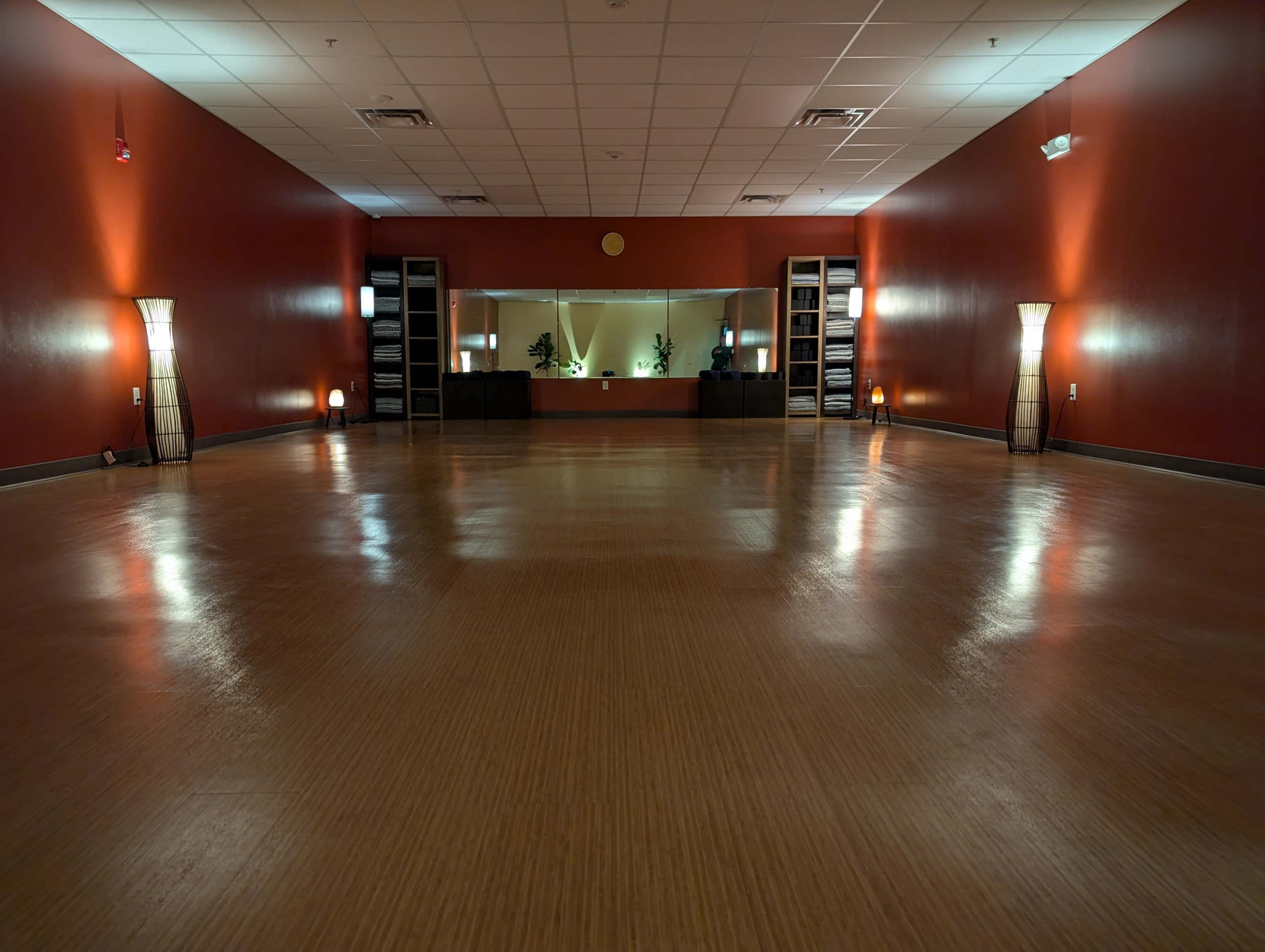 The image shows a spacious room with a polished wooden floor, red walls, and soft lighting from floor lamps, featuring a mirrored wall at one end.