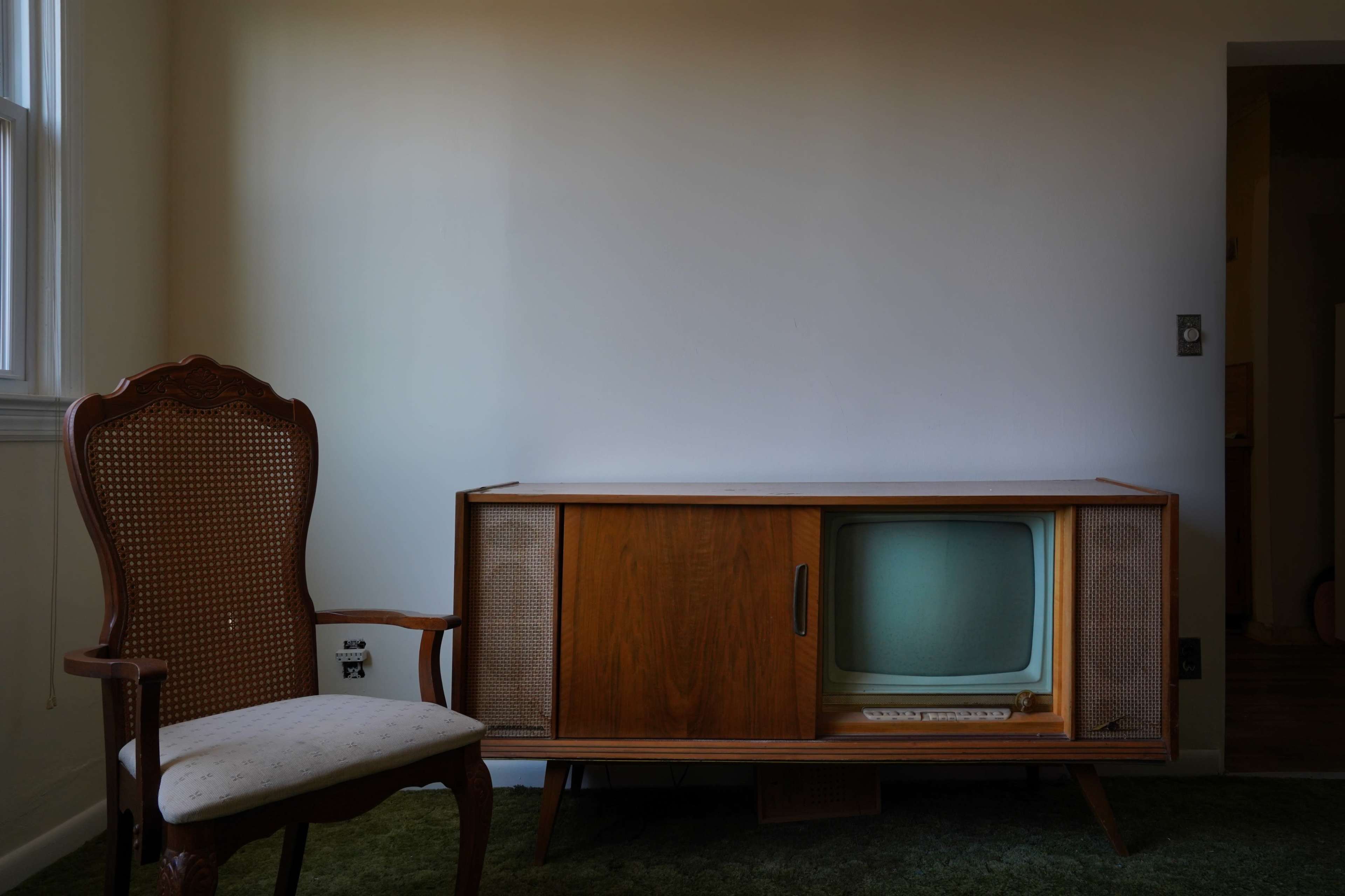 A vintage wooden television set is positioned next to an ornate upholstered chair in a sparsely furnished room.
