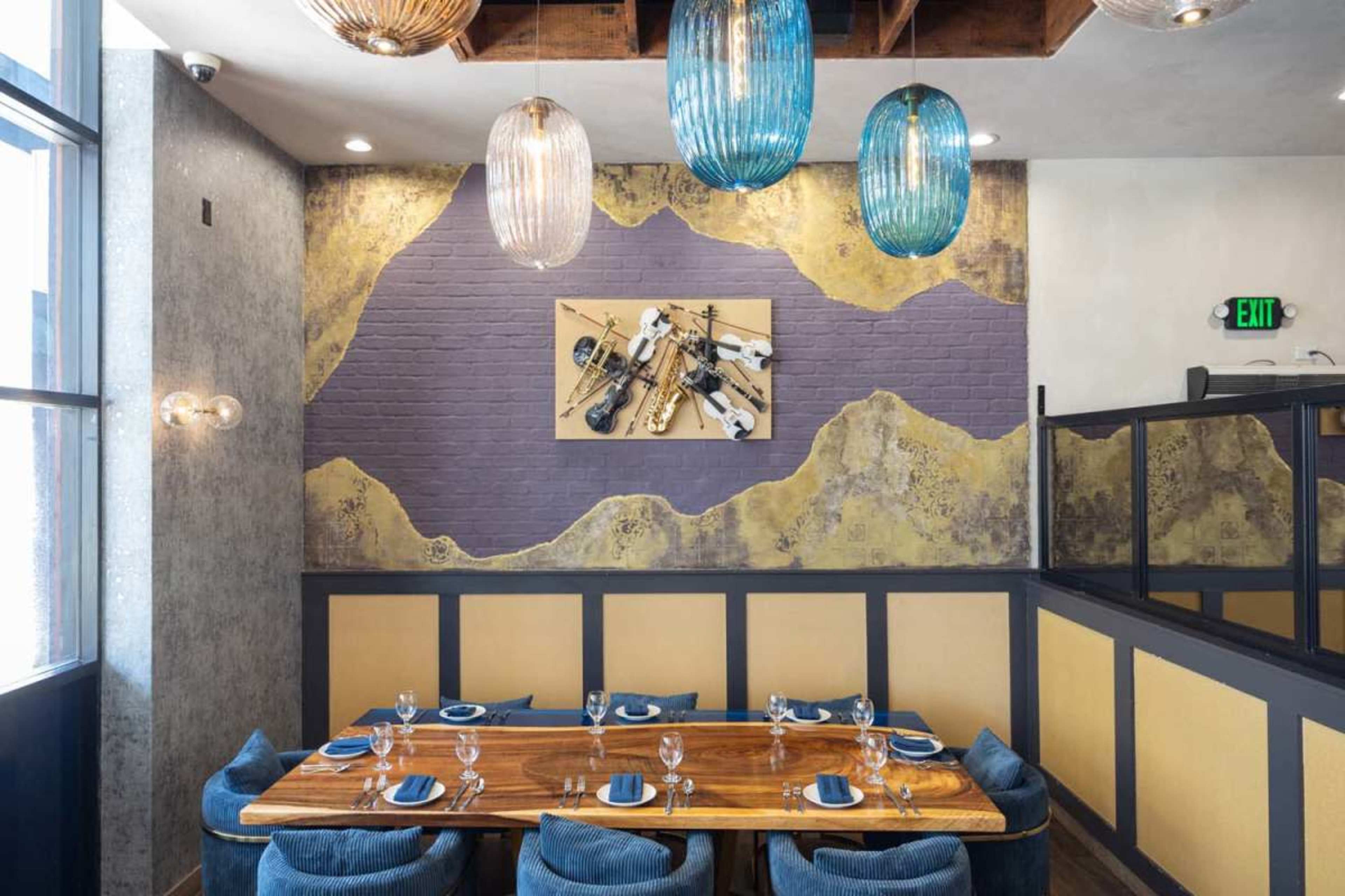 The image shows a modern restaurant interior featuring a wooden table set with blue chairs, a large artwork of musical instruments on the wall, and decorative pendant lights overhead.