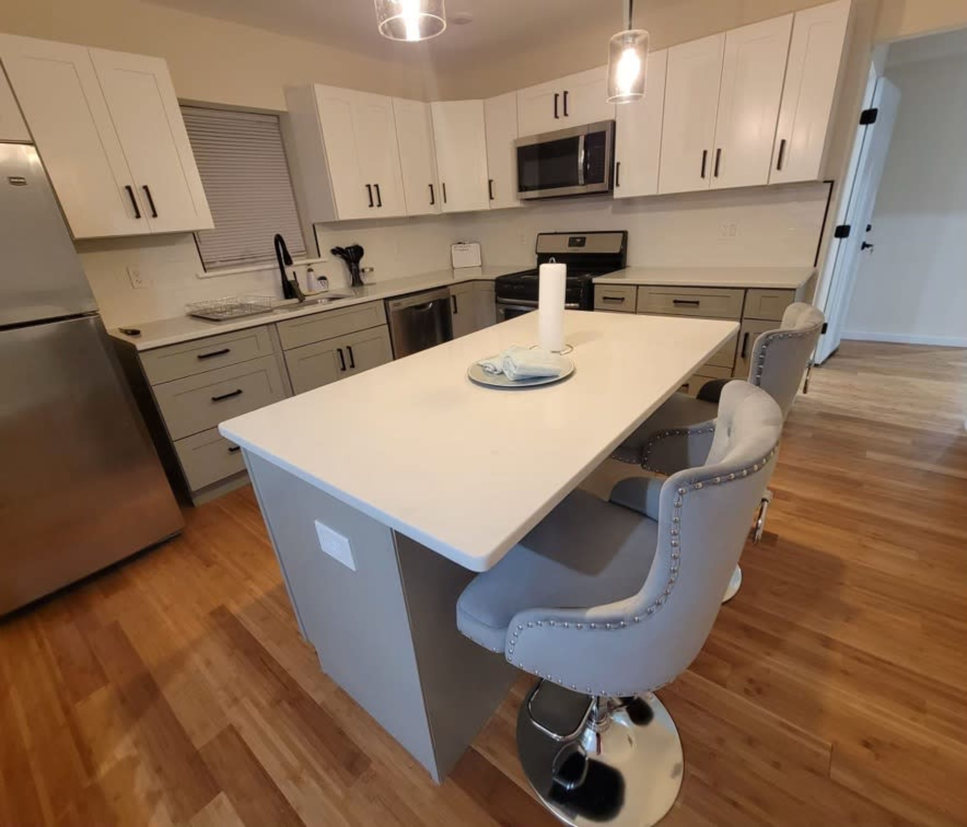 The image shows a modern kitchen with a central island, gray cabinetry, stainless steel appliances, and four upholstered counter stools.