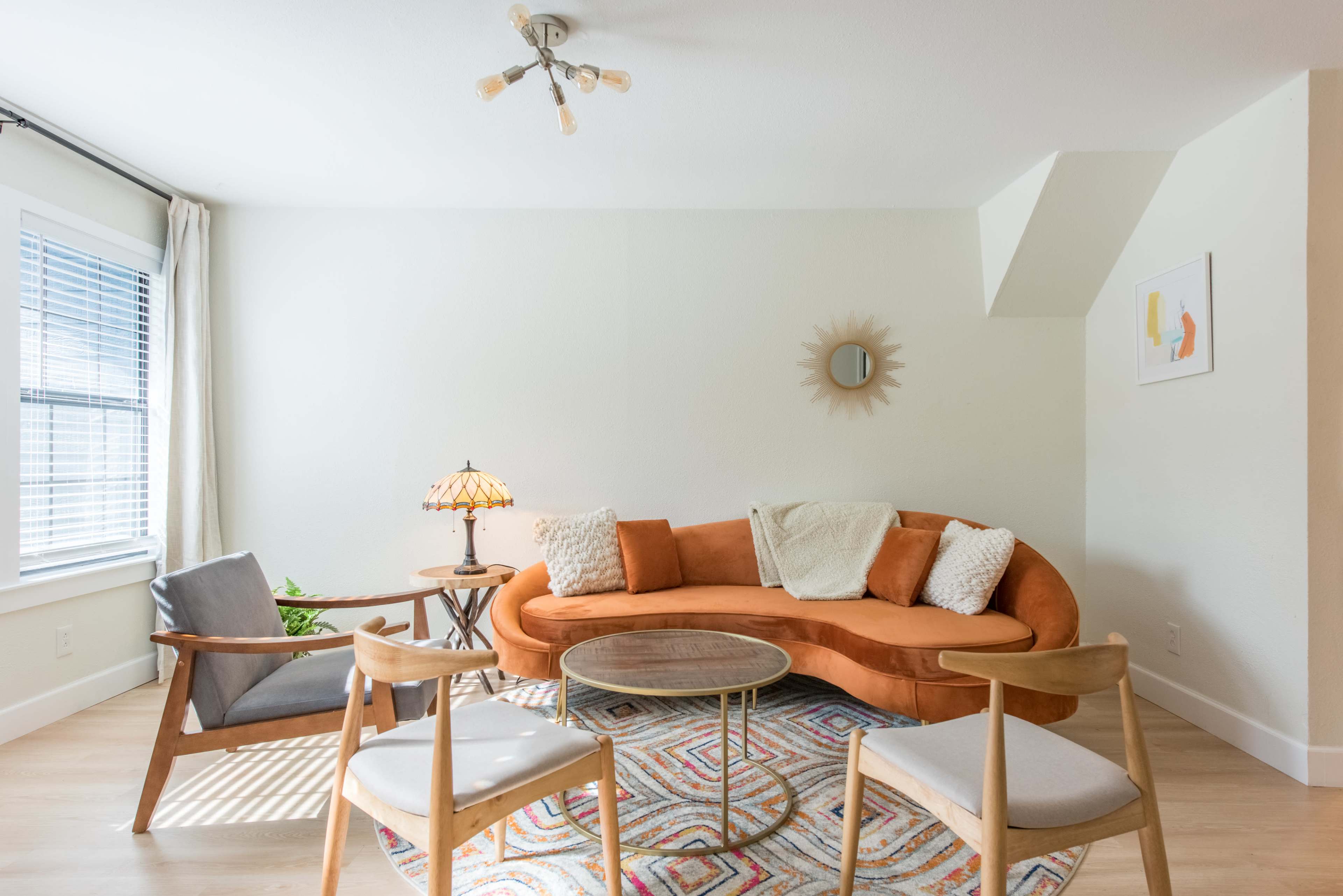 A modern living room features a curved orange sofa, a lamp, a round coffee table, and two chairs, complemented by natural light from a window.