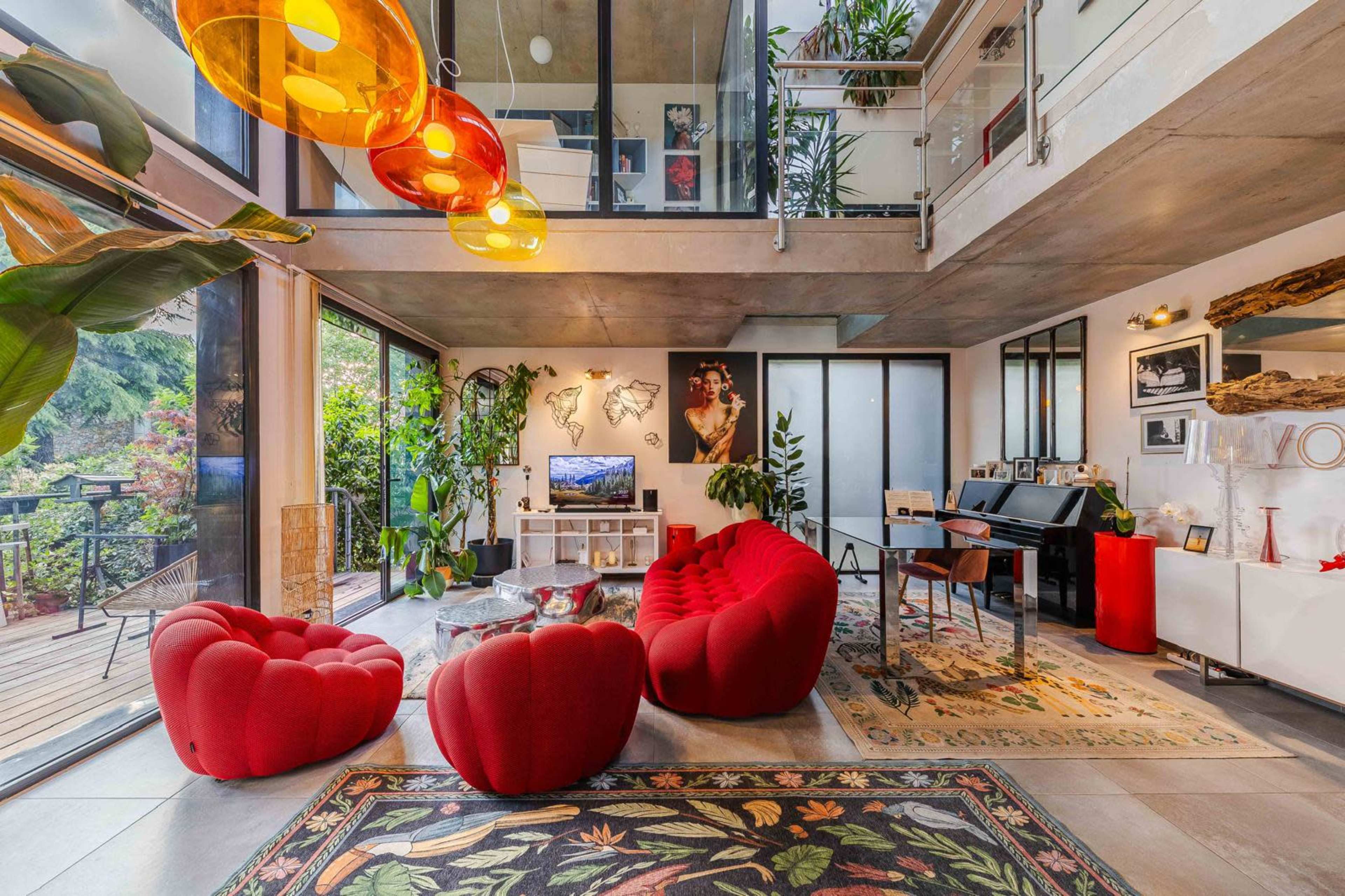 The image shows a modern living space with large windows, featuring a red seating arrangement, a piano, and various plants.