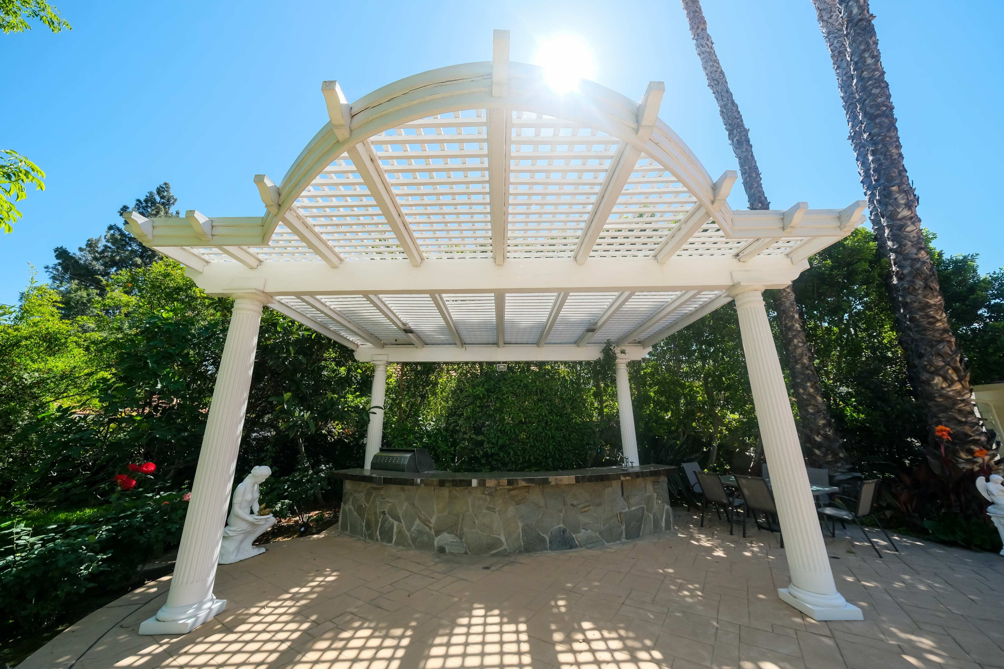 A white pergola with a slatted roof stands over a stone patio surrounded by greenery and palm trees.