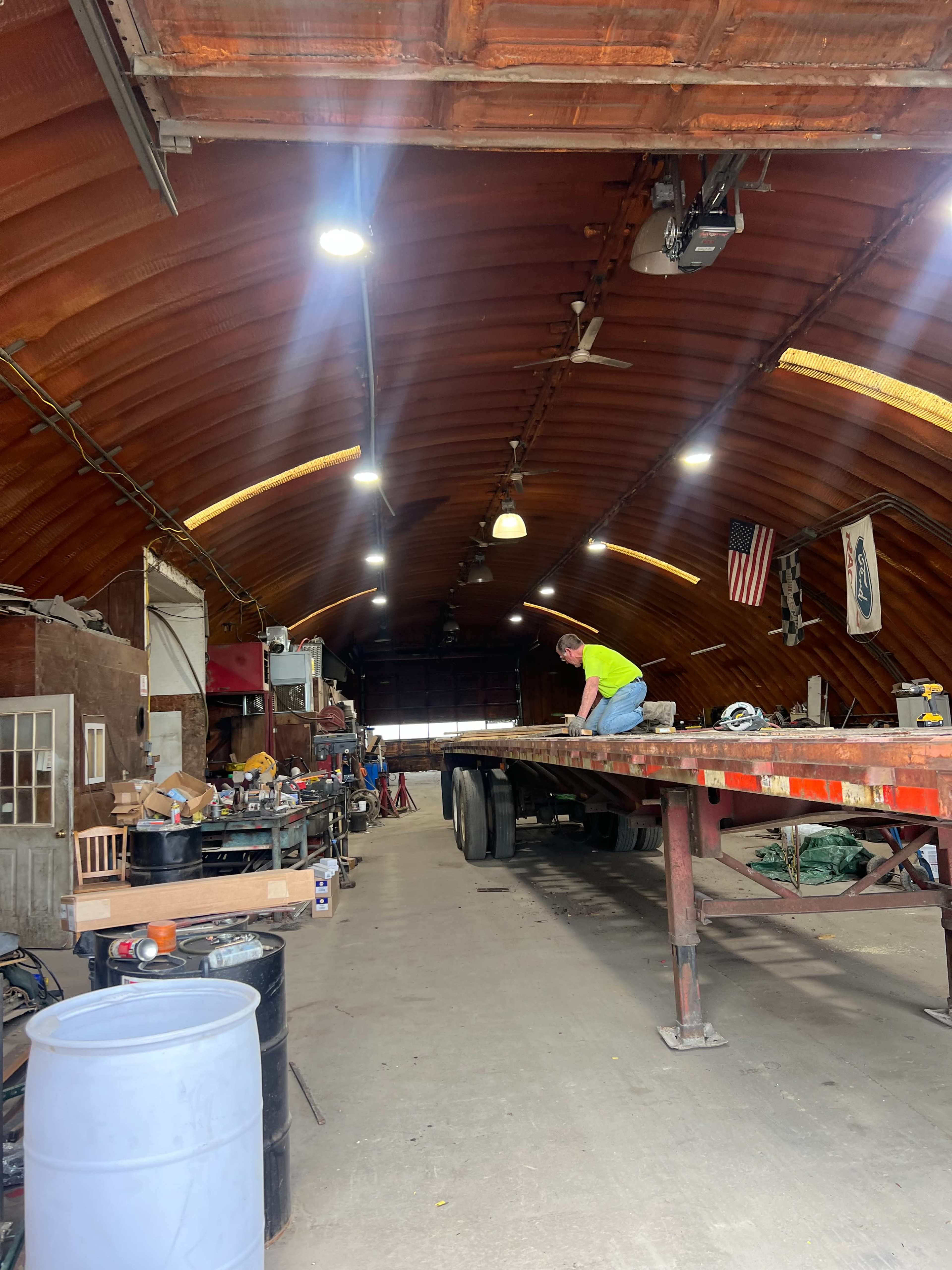 A man works on a flatbed trailer inside a large, well-lit barn garage filled with various tools and equipment.
