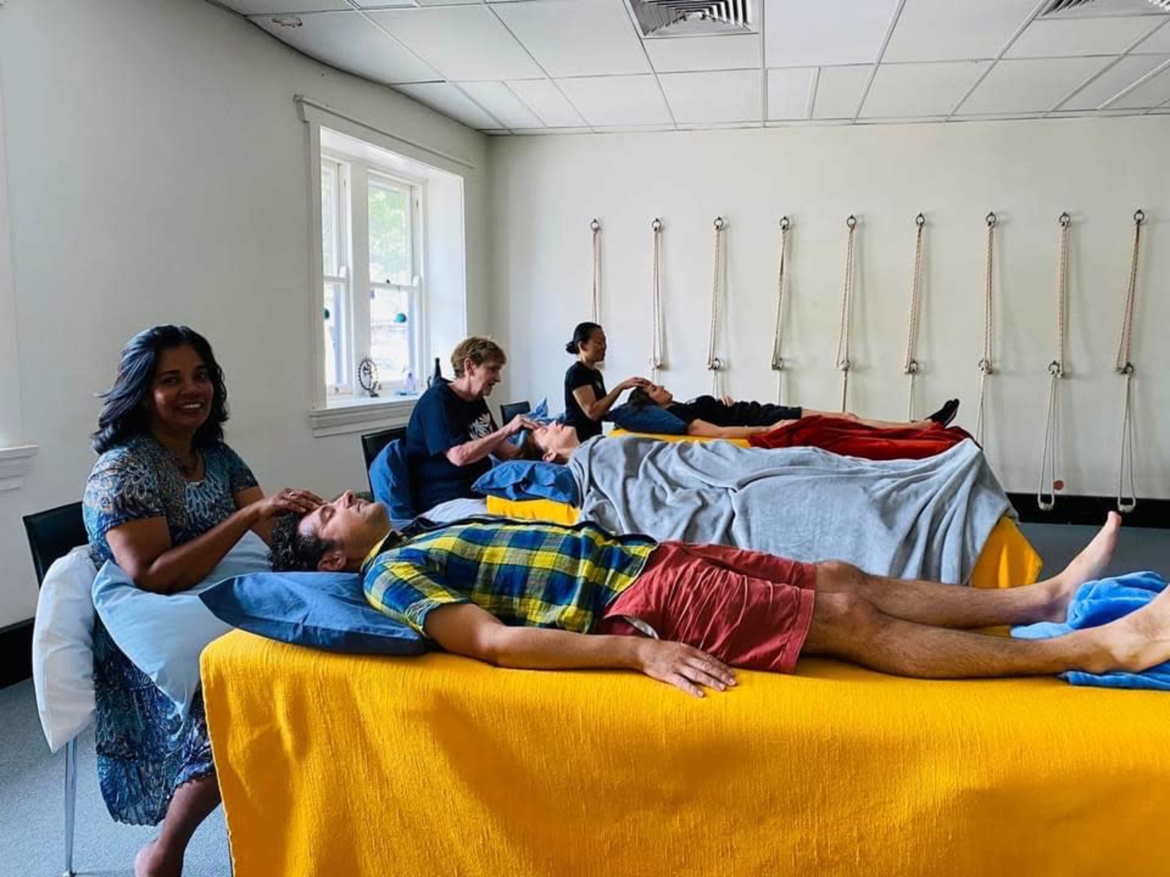 A group of five people is engaged in relaxation activities in a room, with two individuals receiving massage on treatment tables while others sit nearby.