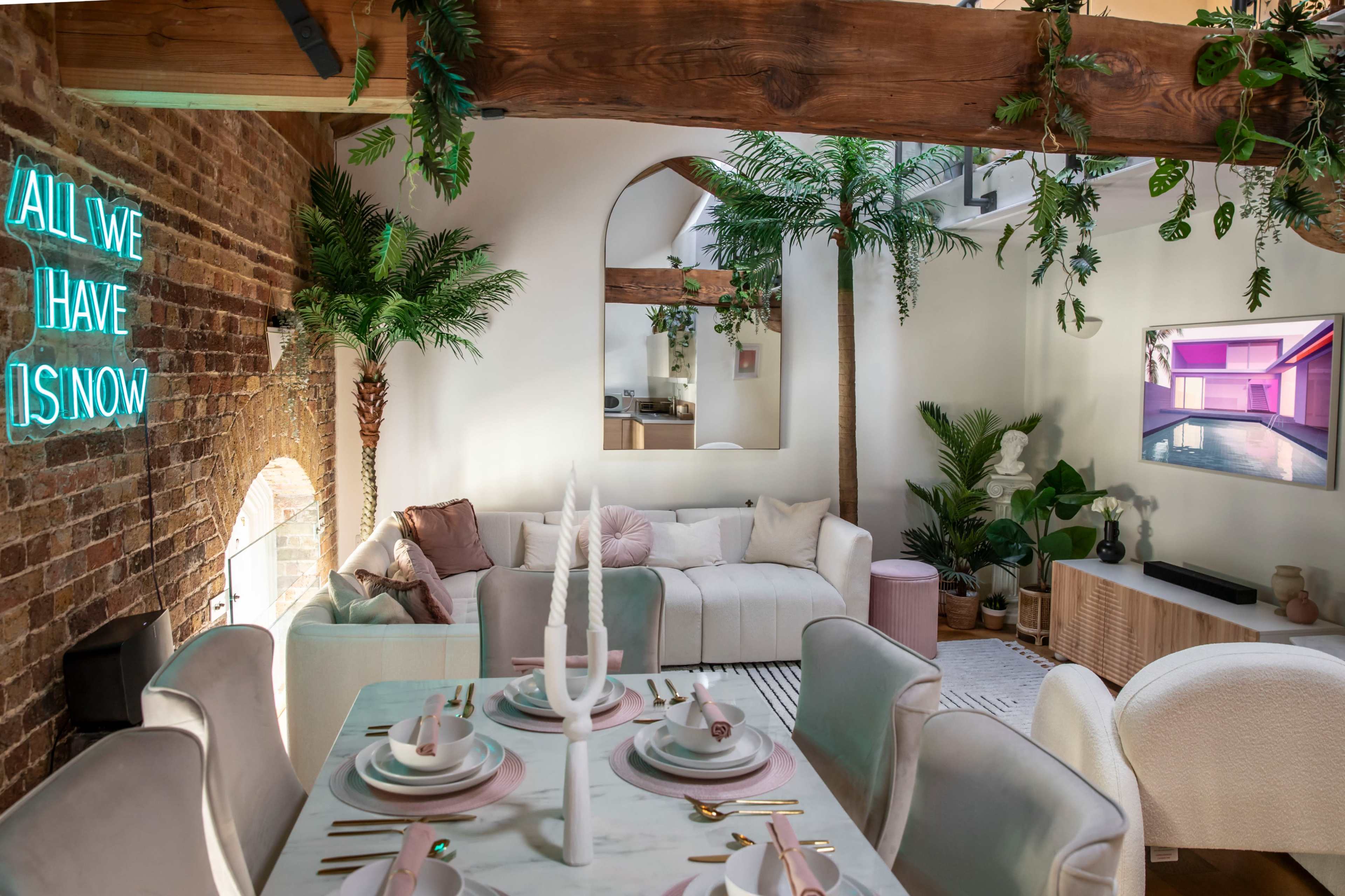 A contemporary living space features a dining table set with elegant tableware, surrounded by greenery and a cozy seating area, highlighted by neon artwork on a brick wall.
