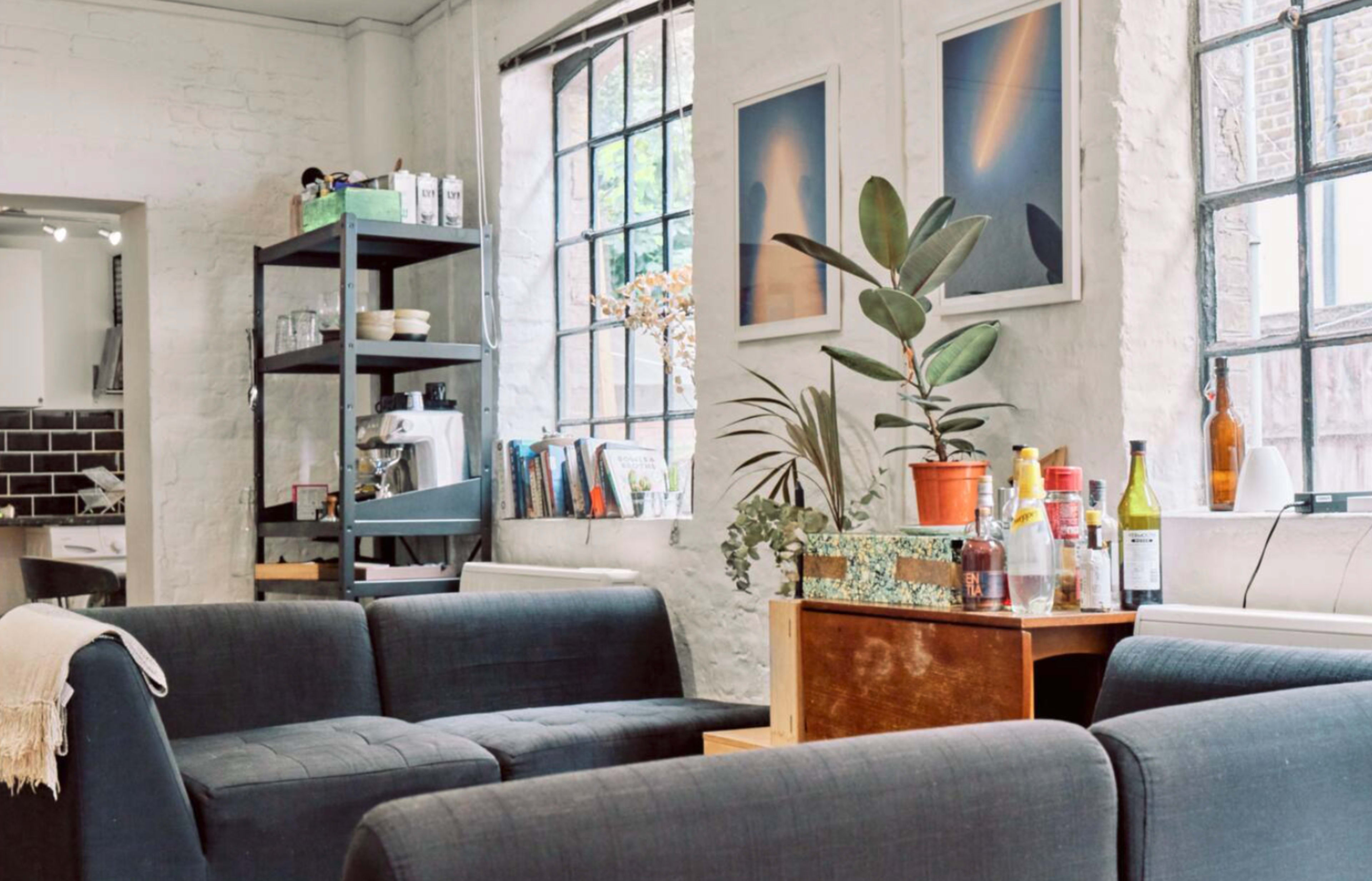 A cozy living space features a black couch arrangement, a wooden coffee table with plants and bottles, and large windows allowing natural light to enter.
