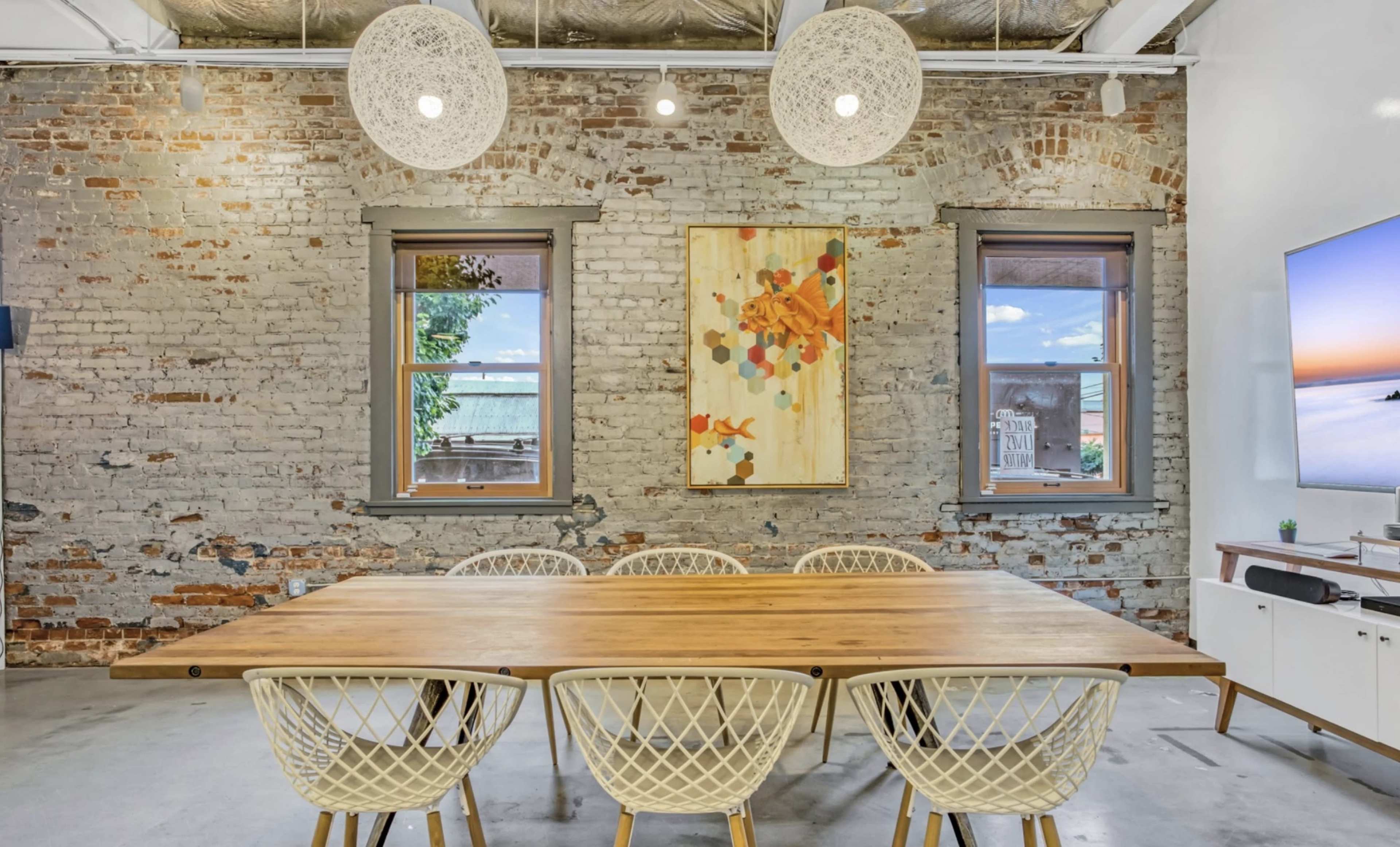 A wooden dining table is centered in a room with exposed brick walls and large windows, illuminated by two pendant lights.