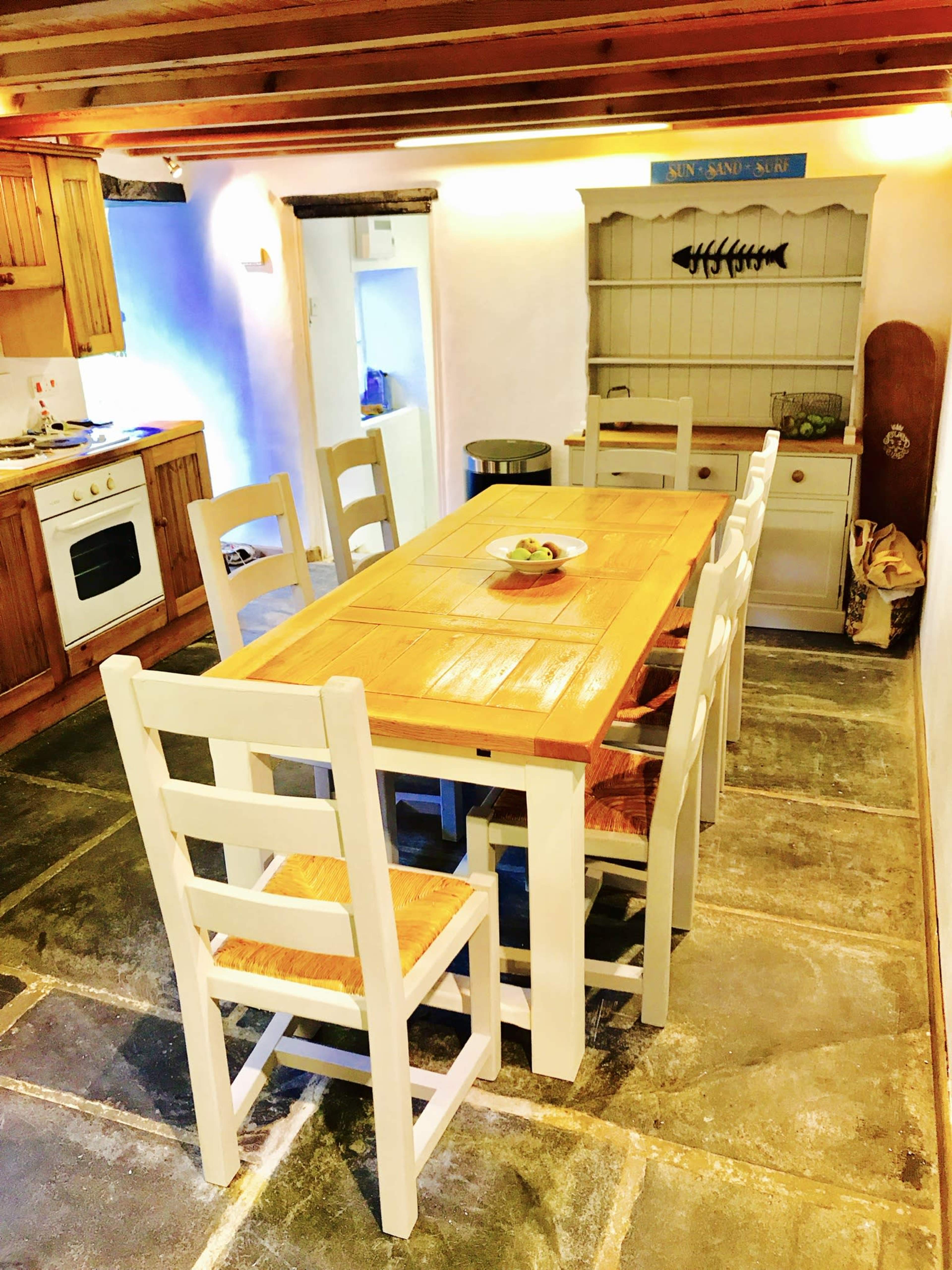 The image shows a wooden dining table surrounded by white chairs in a kitchen with wooden cabinets and a shelf, featuring a bowl of fruit in the center.