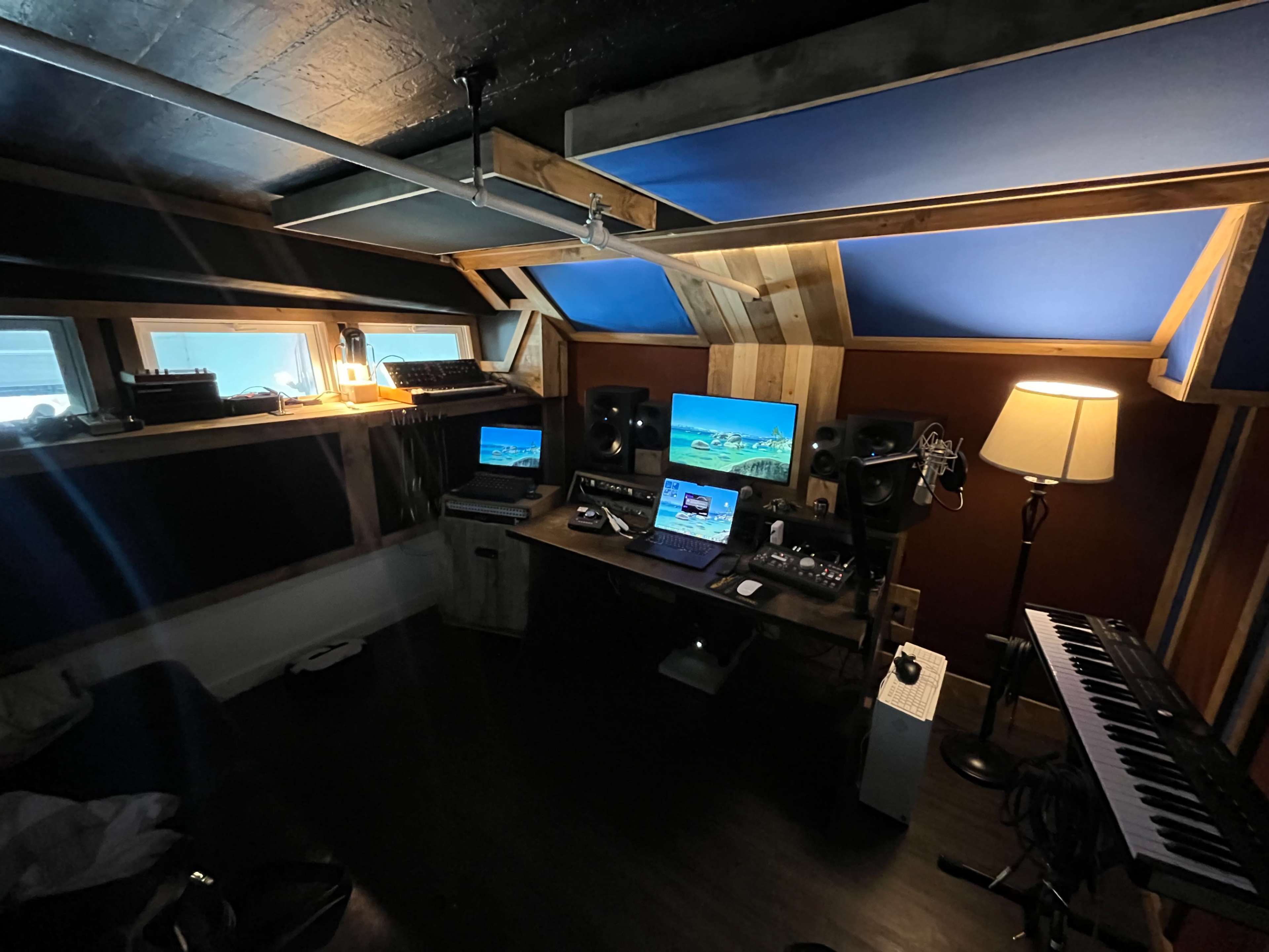 The image shows a music production studio with multiple monitors, audio equipment, and a keyboard, featuring wooden paneling and soft lighting.
