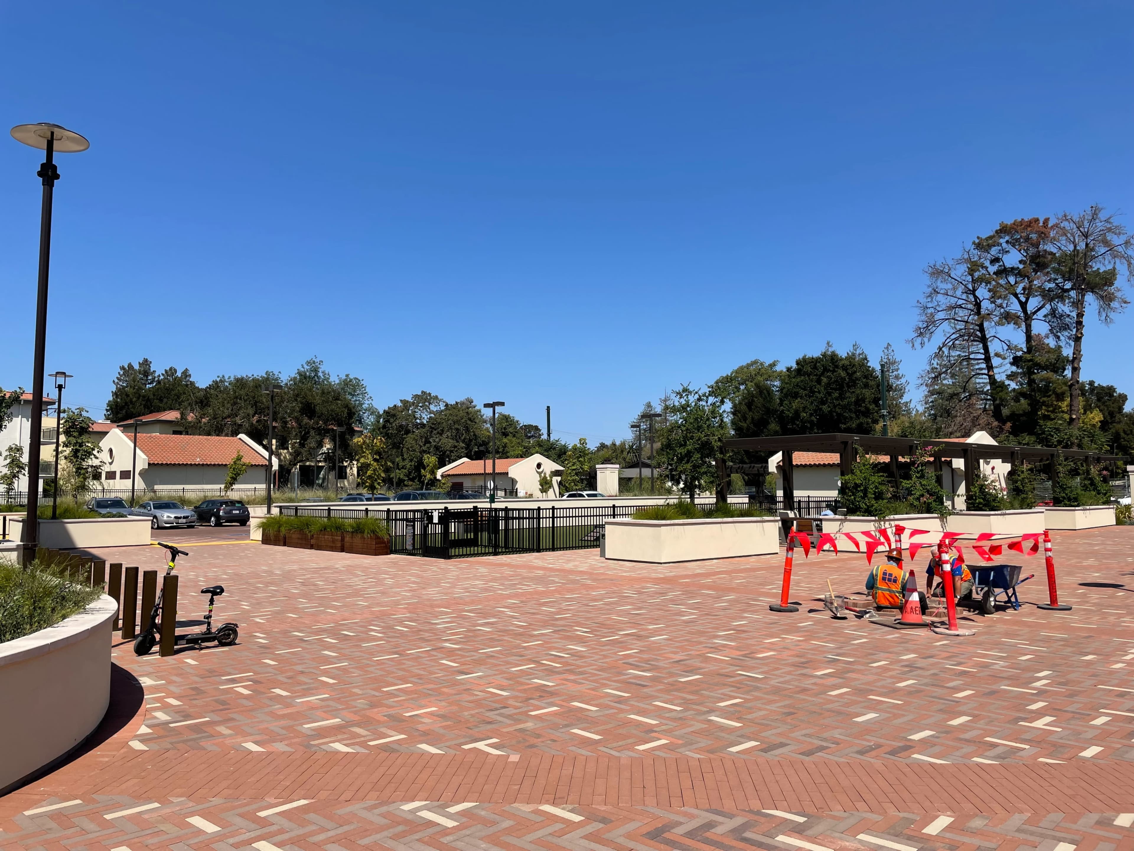 A paved area with construction barriers and equipment in front of a rectangular water feature, surrounded by trees and buildings under a clear blue sky.