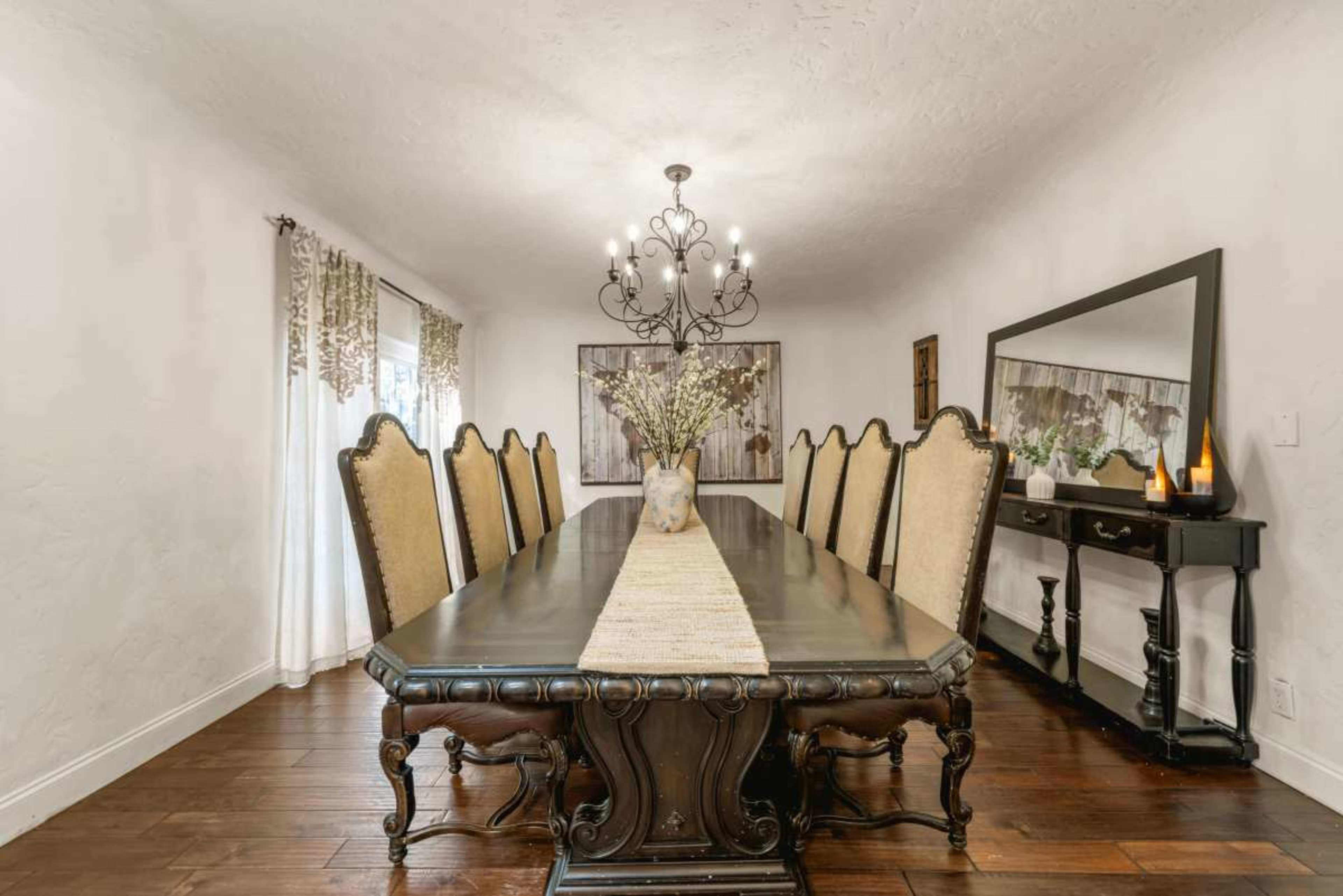 A long dark wooden dining table is set with ten high-backed chairs, under a large chandelier, with a vase of flowers in the center and a mirror along one wall.