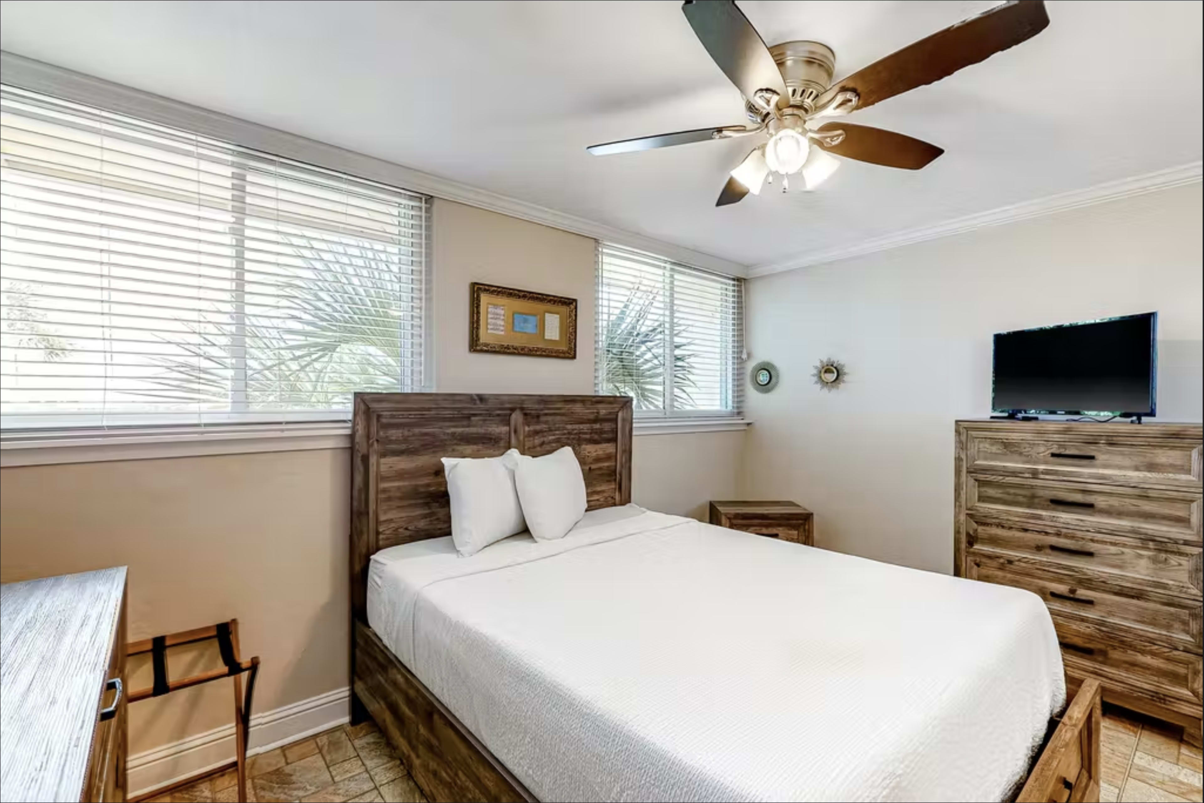 A simple bedroom features a double bed with a wooden headboard, a nightstand, a dresser, and a ceiling fan, with windows providing natural light.