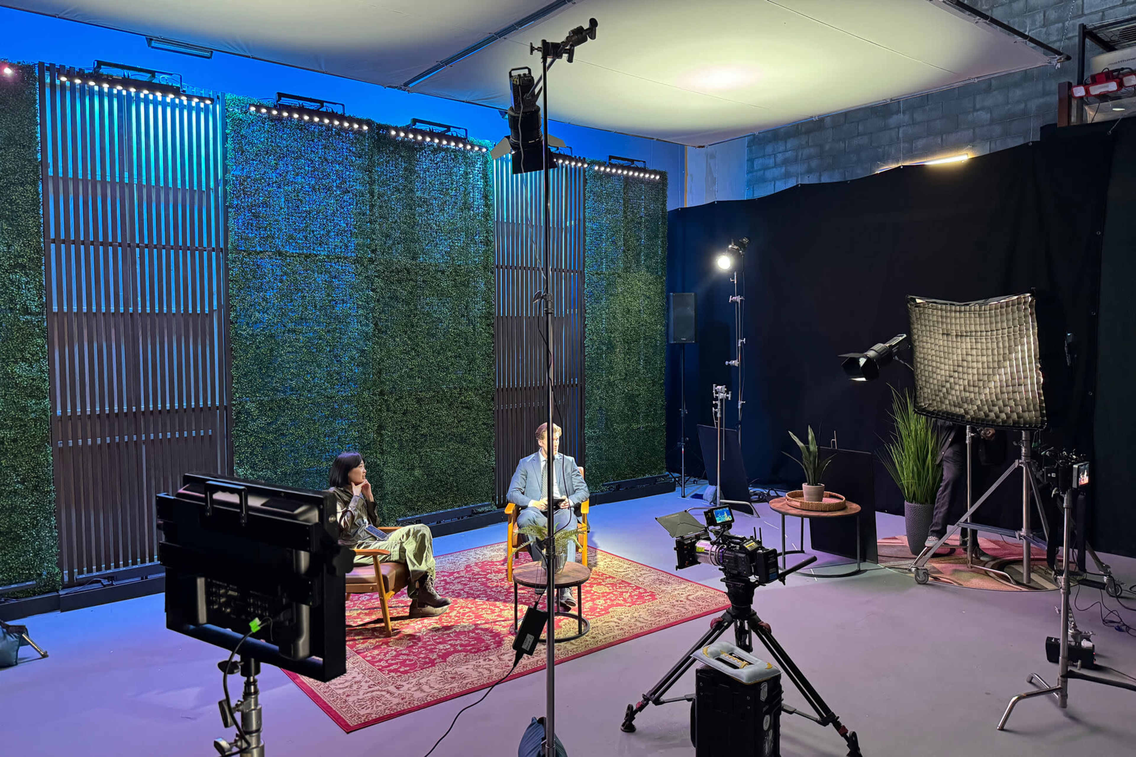 The image shows a studio setup for an interview, featuring two individuals seated on chairs in front of a green backdrop and various lighting and camera equipment.