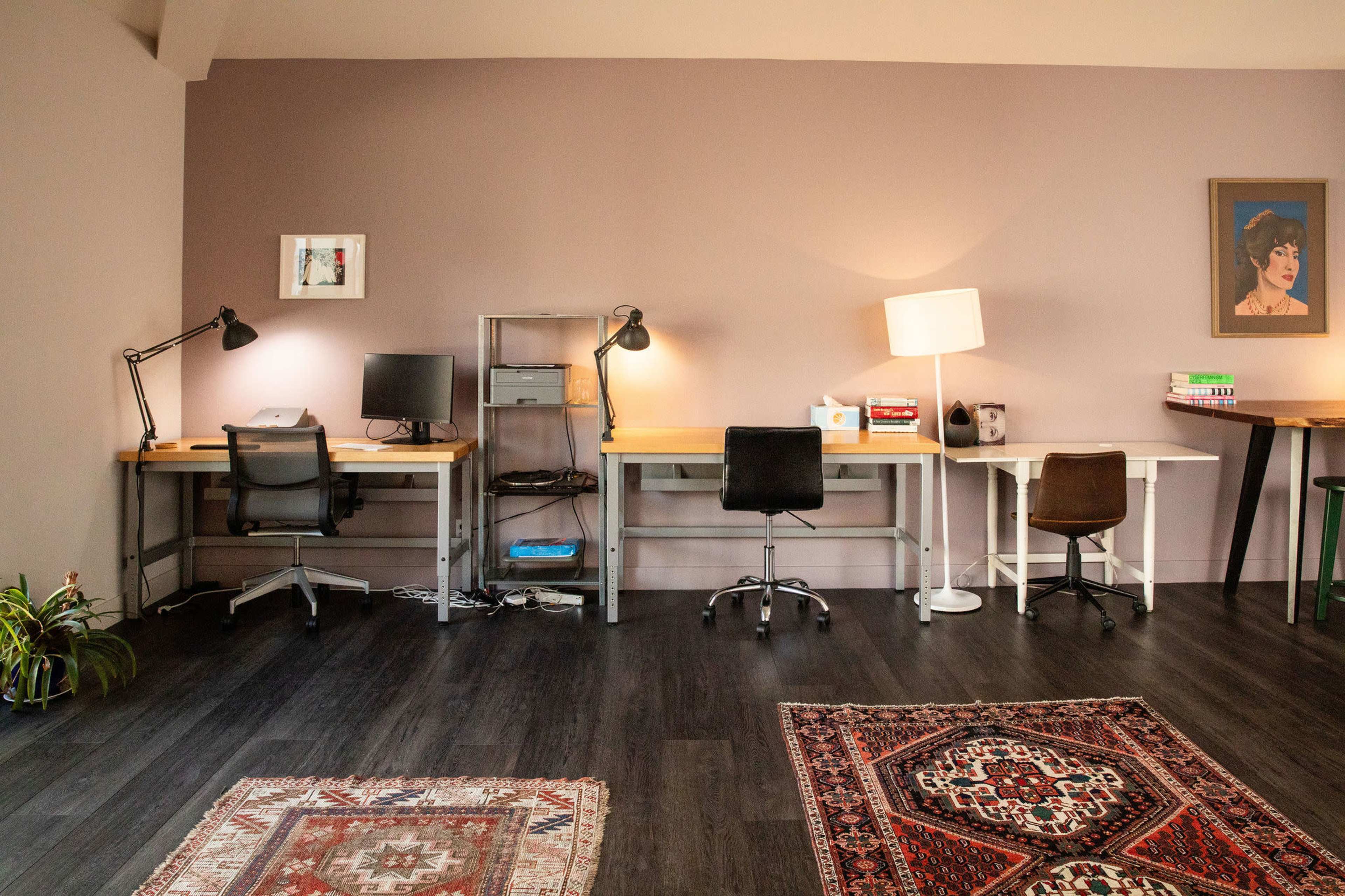The image shows a home office space featuring three desks arranged along a wall, with lamps, a computer, and decorative rugs on a dark floor.