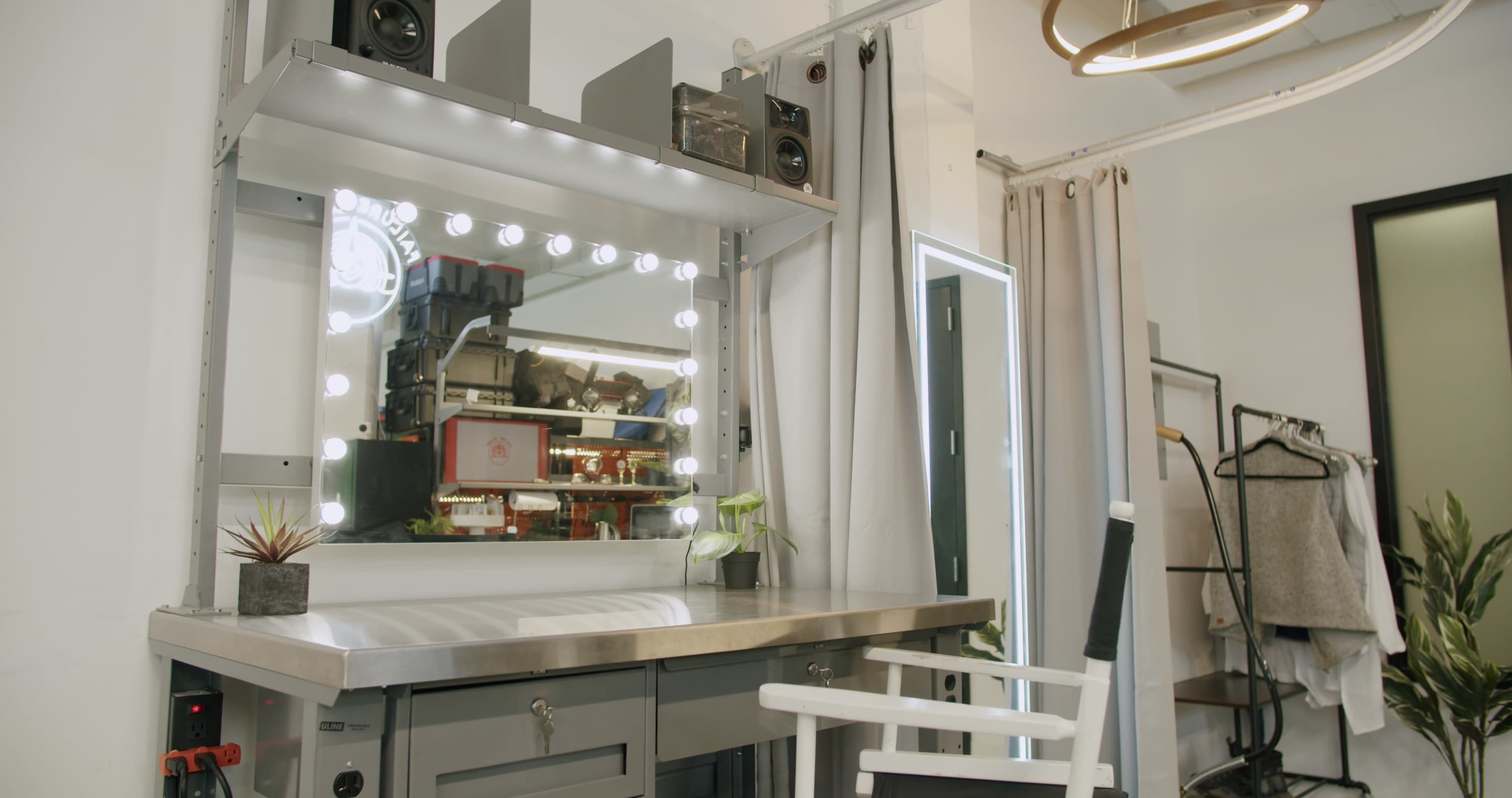 A modern vanity area with a mirror surrounded by lights, a metal countertop, and a white chair next to it.